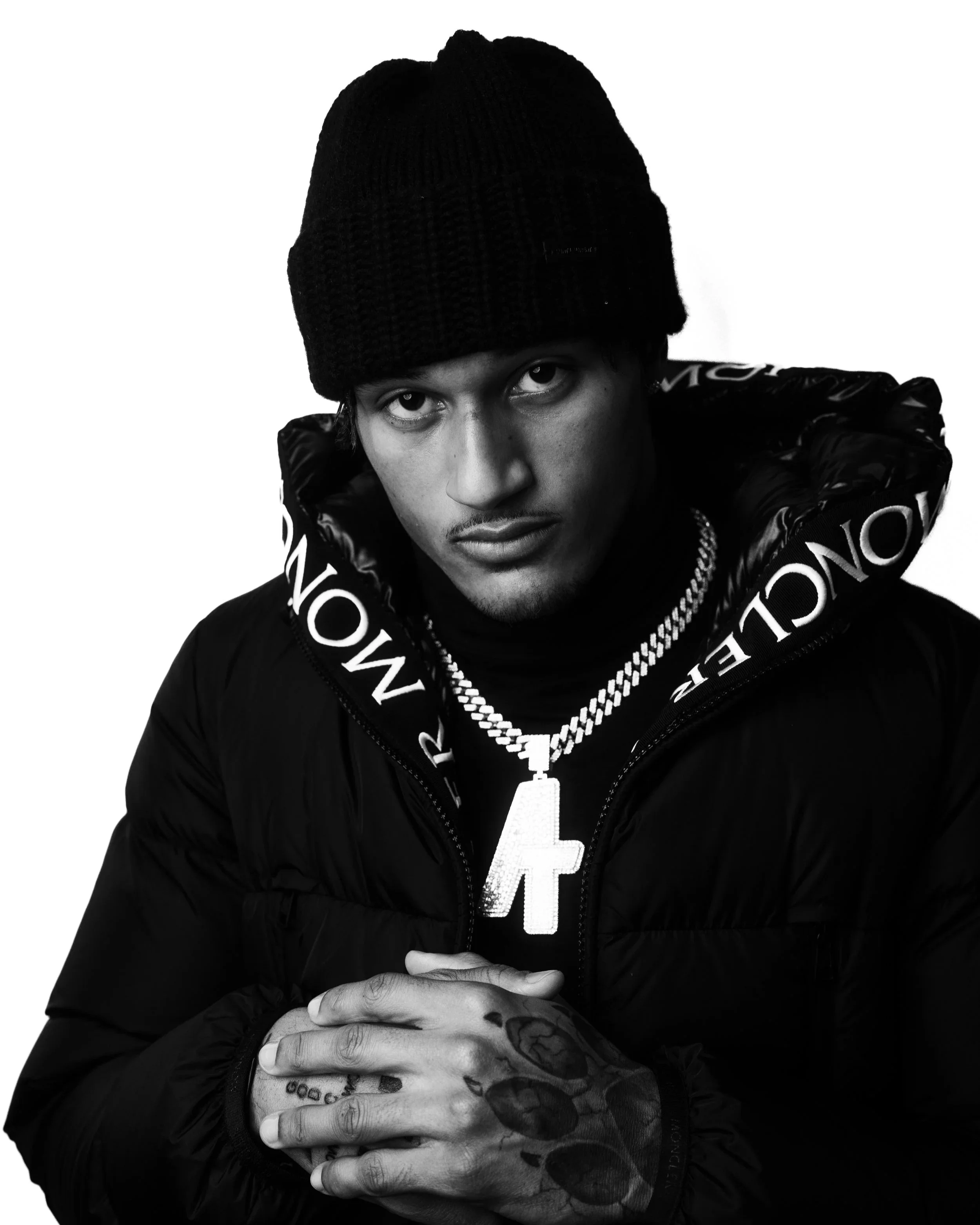 Black and white portrait of Alontae Taylor wearing a black knit beanie and a black jacket with Louis Vuitton branding, posing with his hands clasped and wearing a large chain with a cross pendant, against a white background.