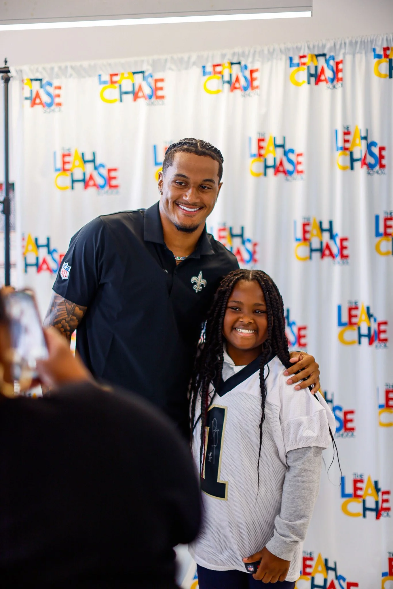 Alontae Taylor in a black Saints collar shirt standing next to a young girl in a white sports jersey, both smiling, at an event with a colorful backdrop reading 'LEAP LEAD LEAD'.