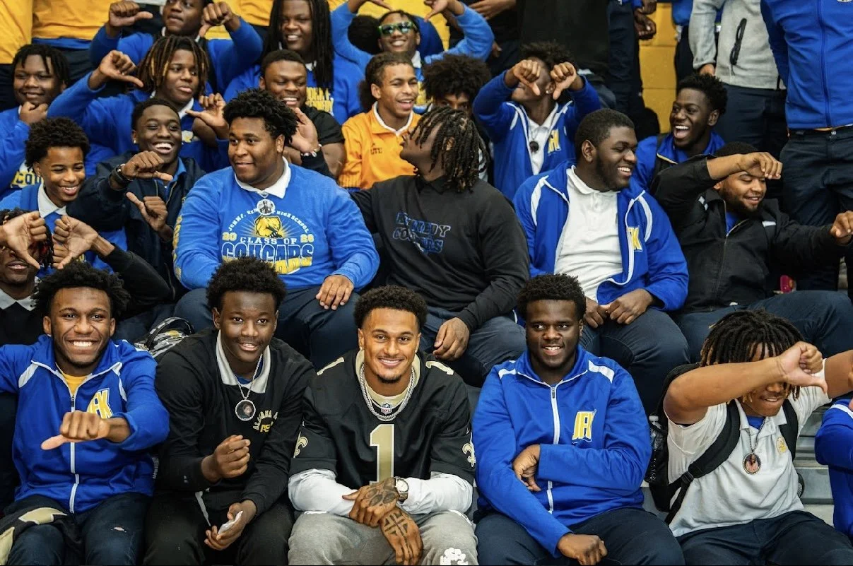 Group of high school students and athletes sitting on bleachers, smiling, and flexing their arms, wearing blue and black jackets, some with medals, in a gymnasium.