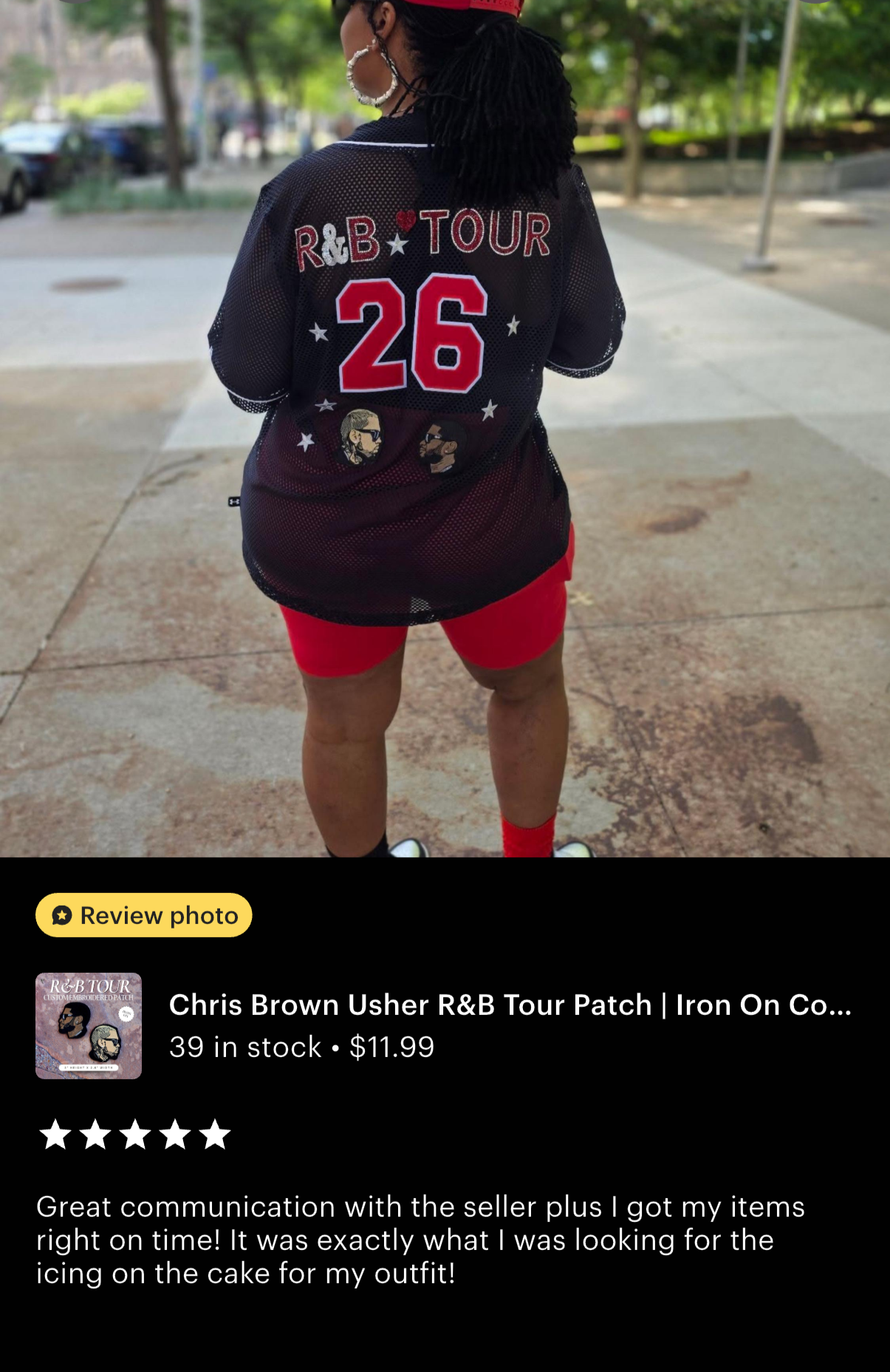 Back view of a woman wearing a black mesh sports jersey with red and white lettering, including the number 26, and illustrations of two faces, standing outdoors on a sidewalk with trees and parked cars in the background.