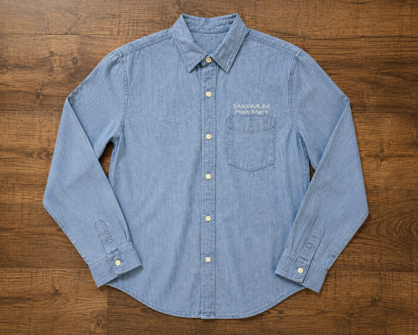 Light blue denim button-up shirt with a front pocket and embroidered text that reads 'MAXIMUM Pest Mgmt' on the left chest, laid flat on a wooden surface.