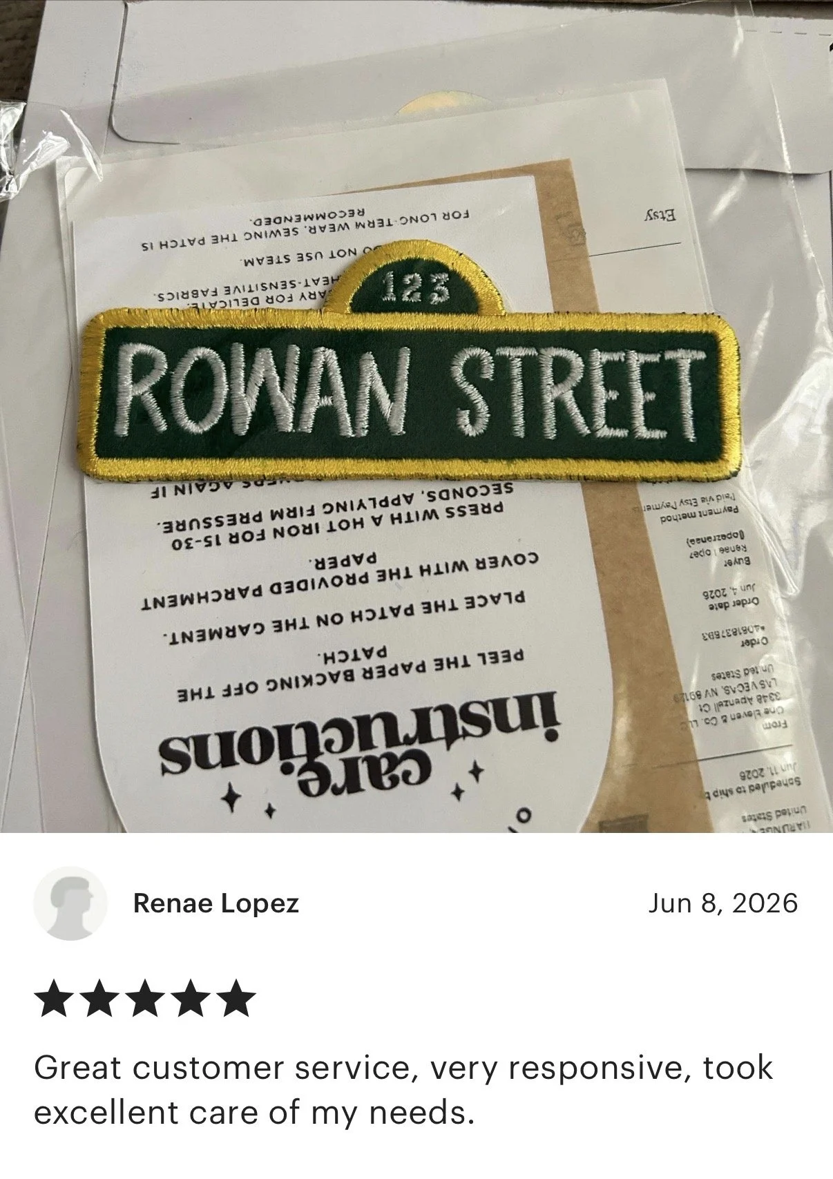 Close-up of a green and yellow embroidered patch with the text '123 ROWAN STREET' and an orange semi-circle at the top, placed on top of a transparent plastic bag containing papers and a receipt.