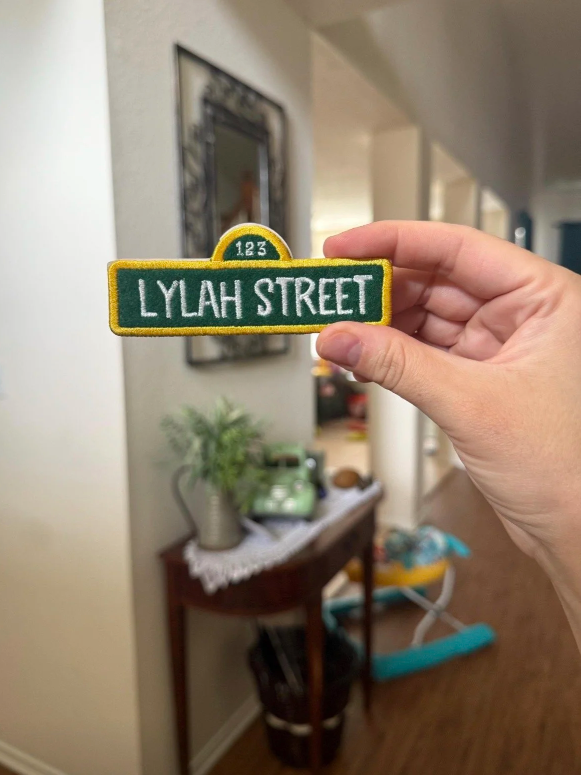 A hand holding a green and yellow embroidered patch with the words 'Lylah Street' and '123' in front of a blurred interior home setting.