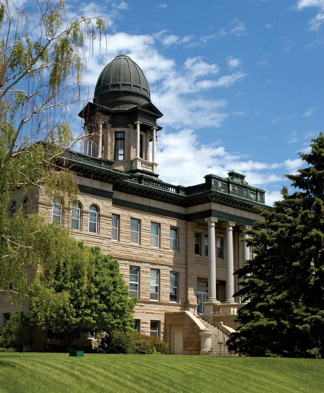 A historic stone building with a large black dome and tall white columns on the front entrance, surrounded by lush green trees and a well-maintained lawn, under a partly cloudy blue sky.