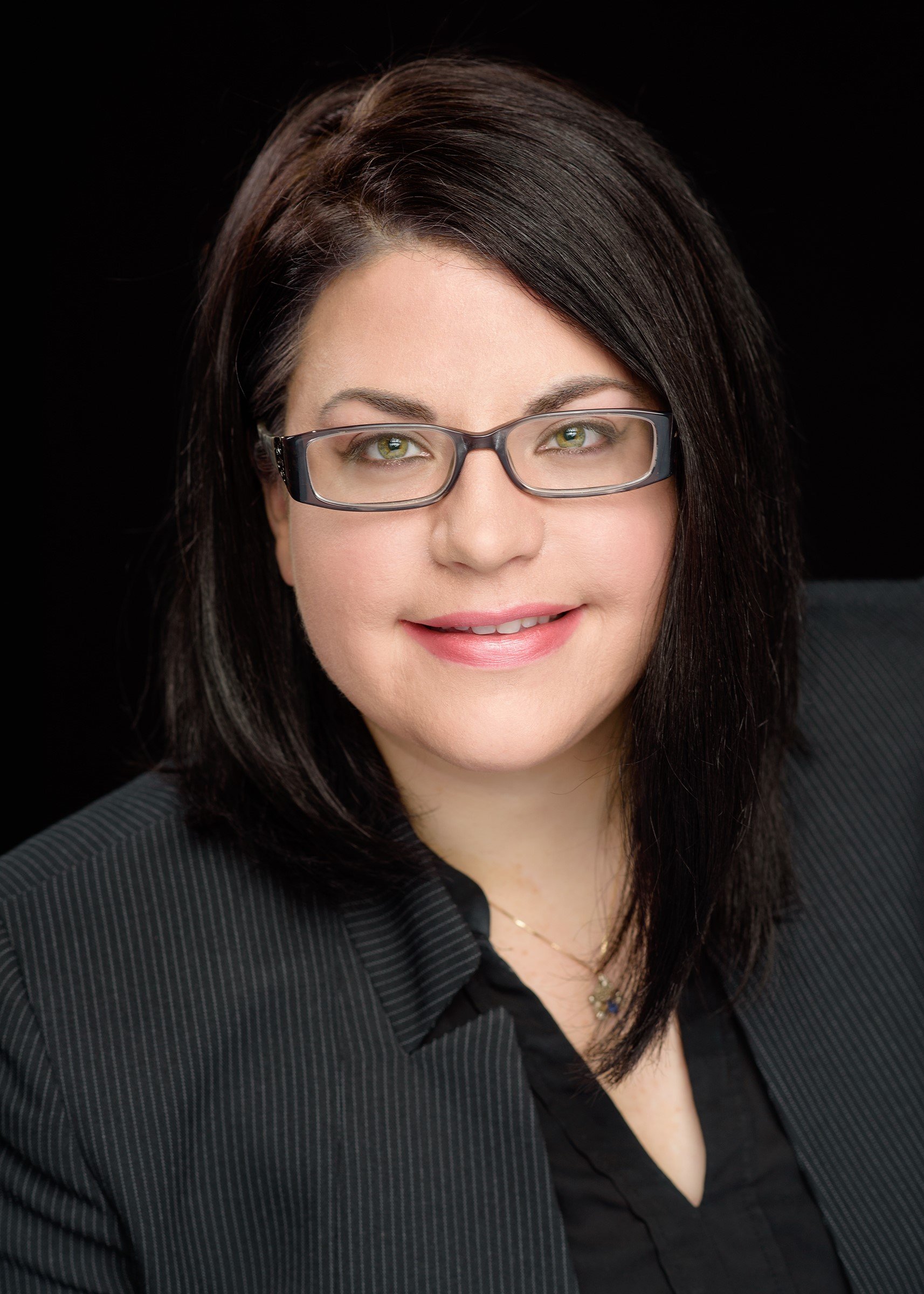 Portrait of a woman with dark brown hair, wearing glasses, a black pinstripe suit, and a black top, smiling against a black background.