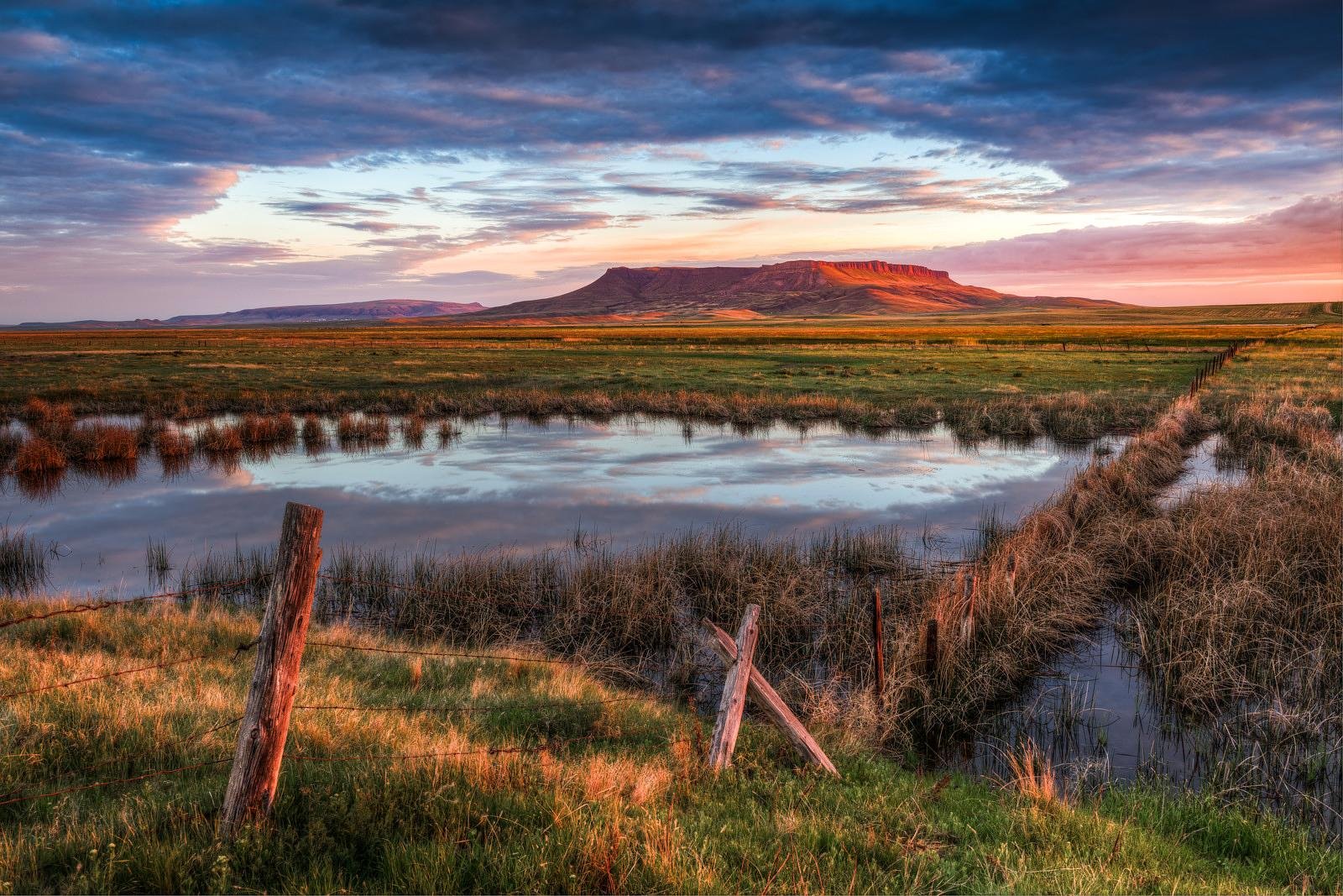A landscape scene at sunset showing a mountain in the background, a cloudy sky, a small pond reflecting the sky and mountain, with grassy fields and a wooden fence in the foreground.
