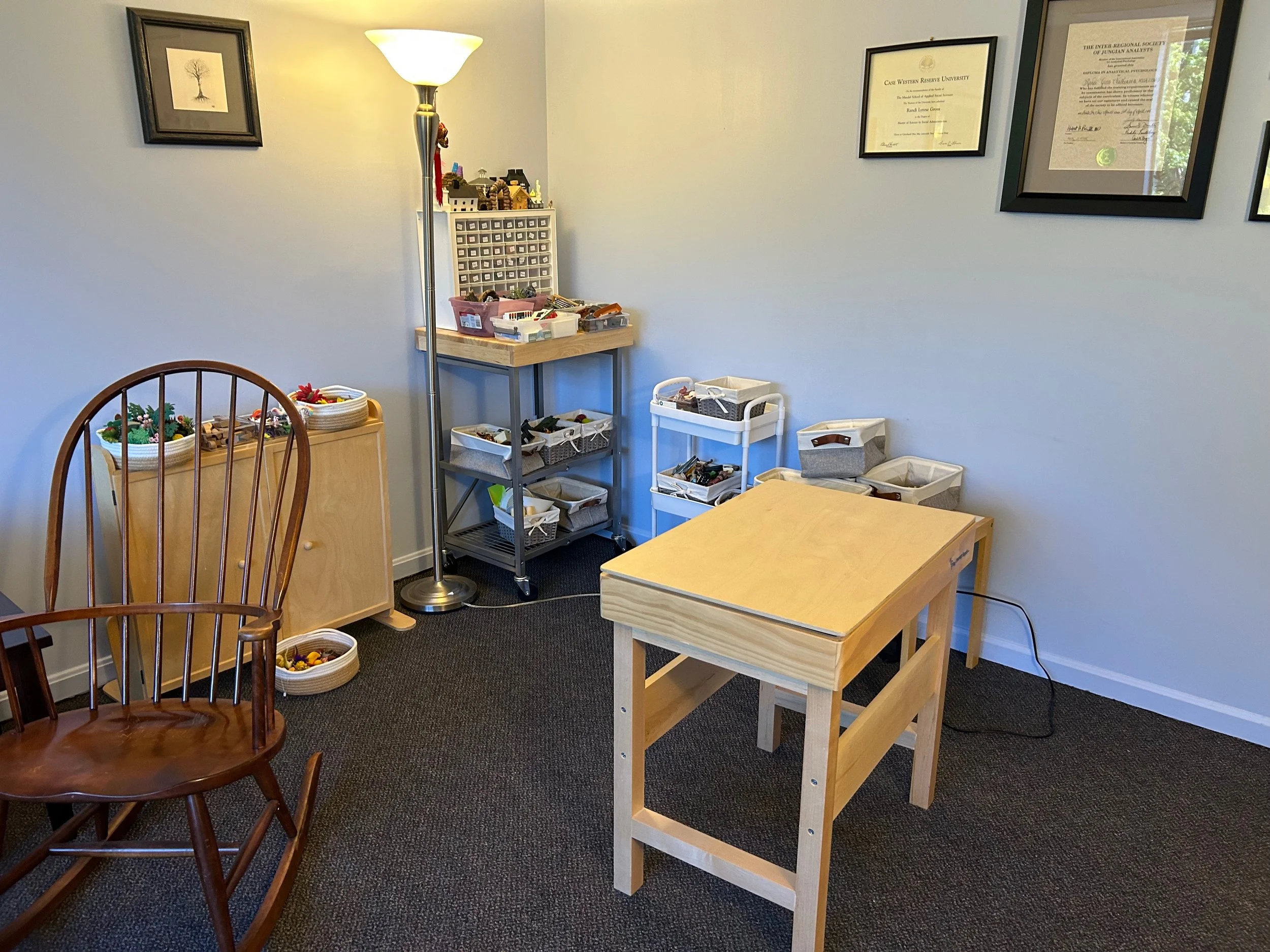 A corner of a room with four framed certificates on the wall, a white lamp, a wooden chair, and various storage containers with small items, including a toy set on a small cart.