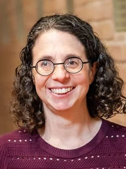A woman with curly dark hair, glasses, and a purple sweater smiling indoors with a blurred brick background.