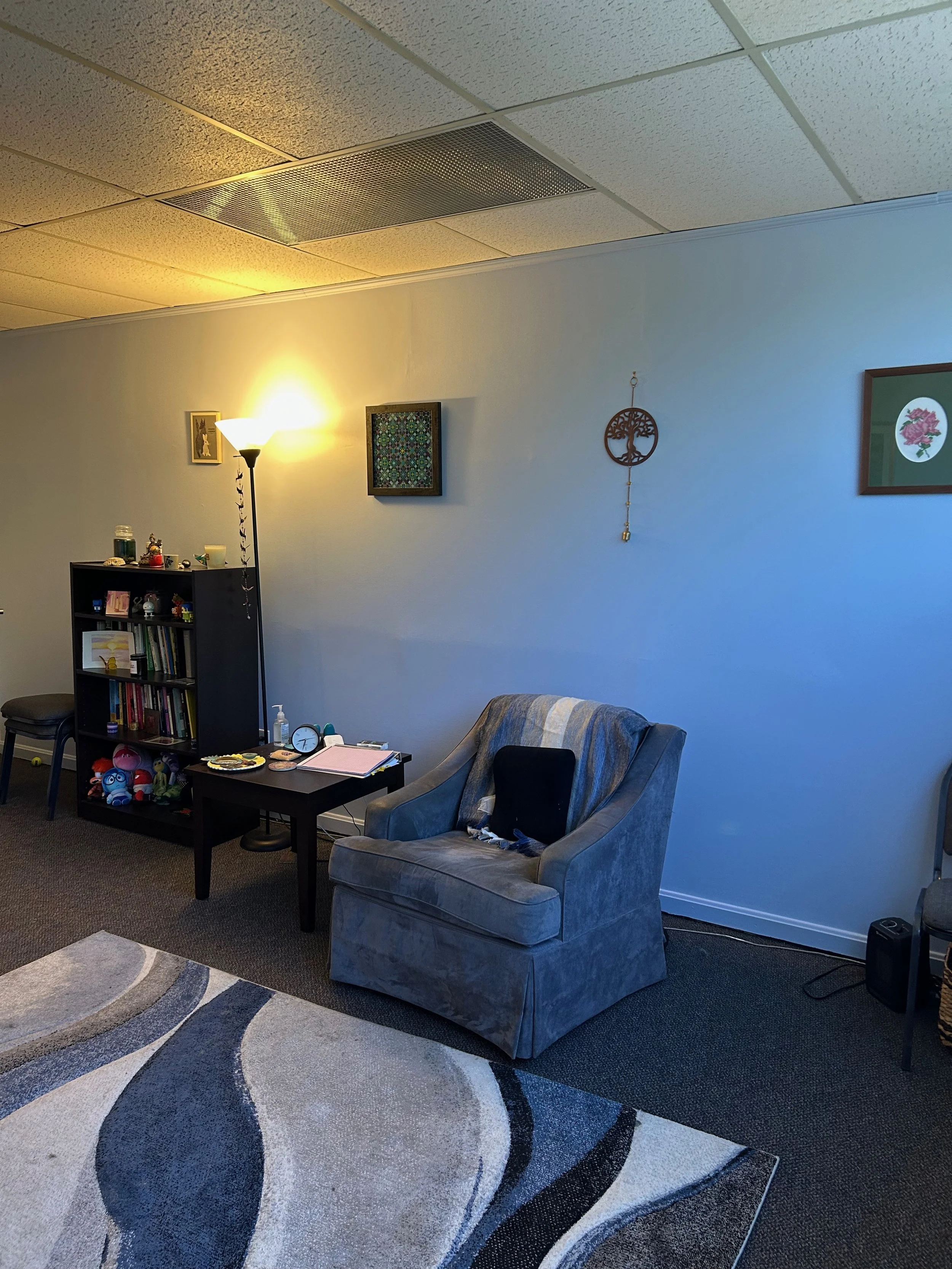 Living room with a gray armchair, a small black side table with jewelry and notebooks, a black bookshelf filled with books and toys, a floor lamp with a yellow glow, two framed pictures and a hanging decorative ornament on the white wall, a black portable speaker on the dark carpeted floor, and a multicolored patterned area rug.