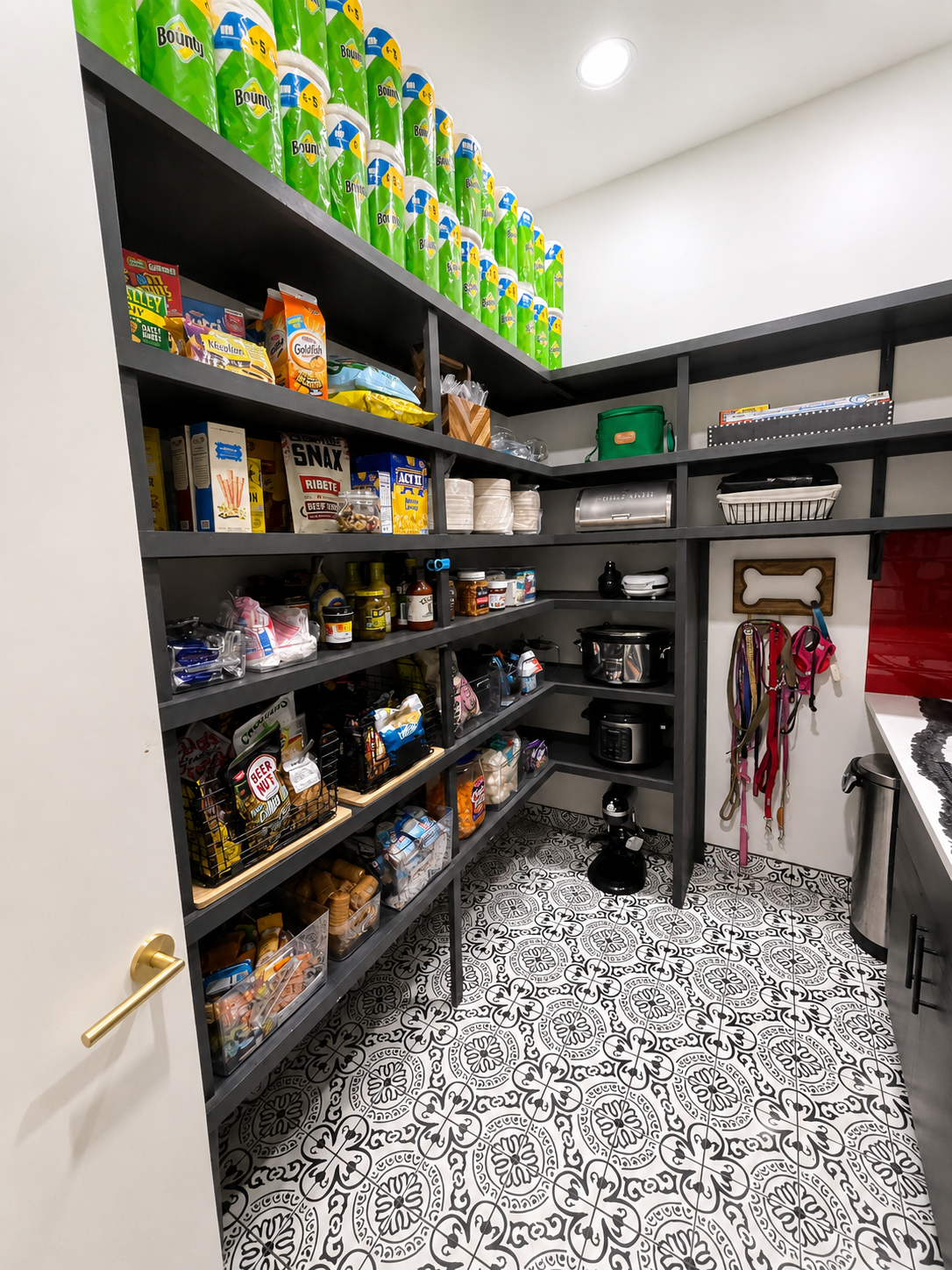 Pantry with shelves filled with household supplies, snacks, and food items; patterned tile floor; dog food containers and appliances visible.
