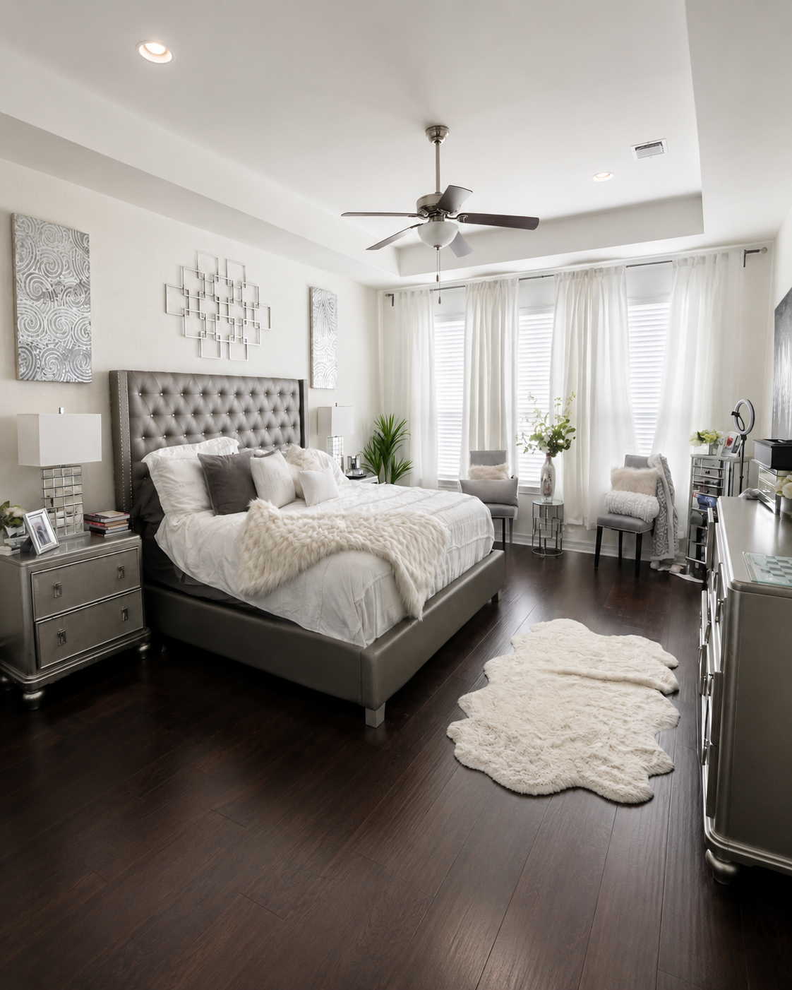 A modern bedroom with a large tufted headboard, white bedding, and dark wood flooring. There are two nightstands with lamps, a small white furry rug, and an arrangement of chairs and decorations near the window, which has white curtains and blinds.