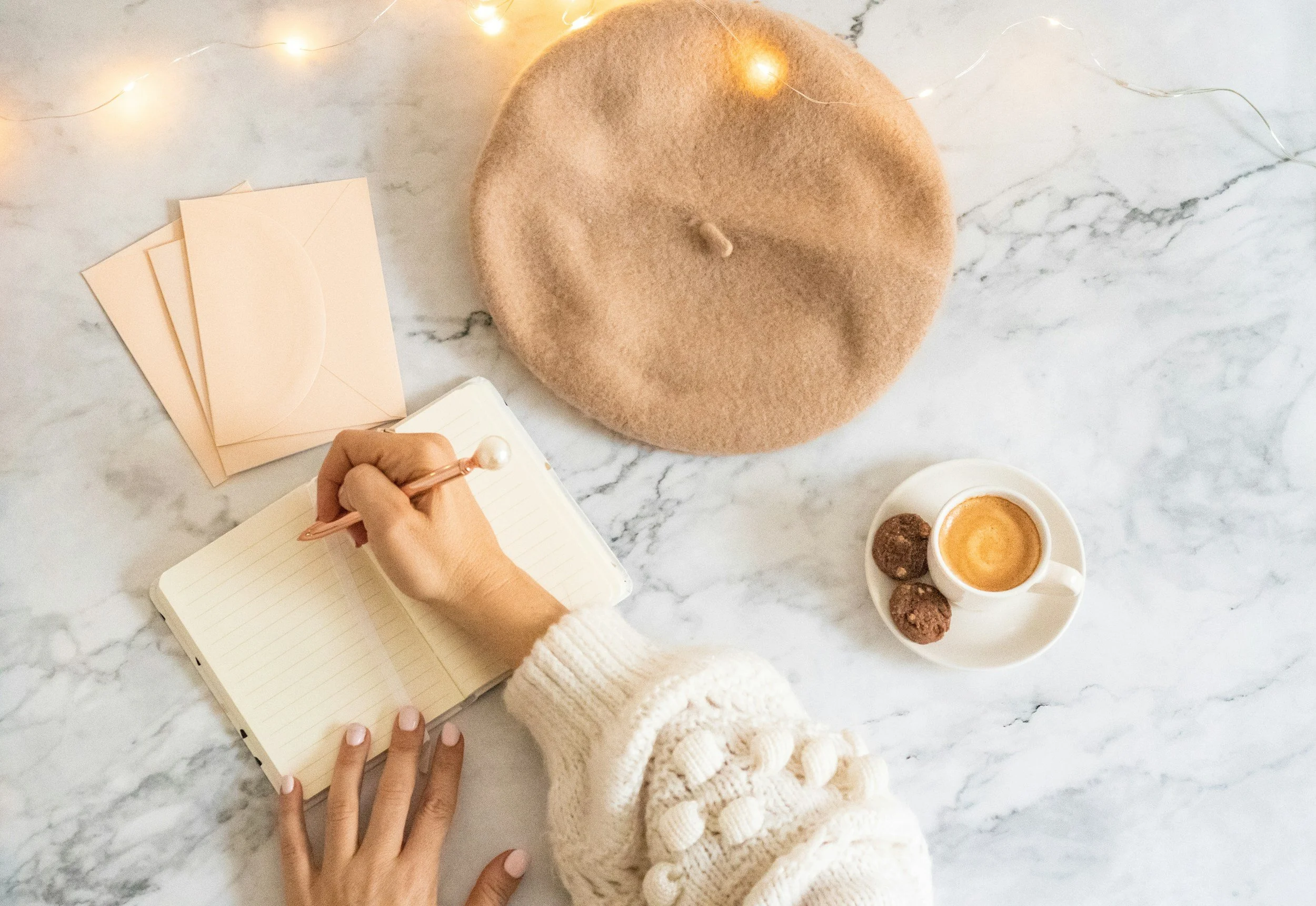 Overhead view of a person's hand writing in a lined notebook with a pink pen, on a marble surface alongside a beige beret, light pink envelopes, a cup of coffee with foam art, and three chocolate or truffle treats, with a string of fairy lights in the background.