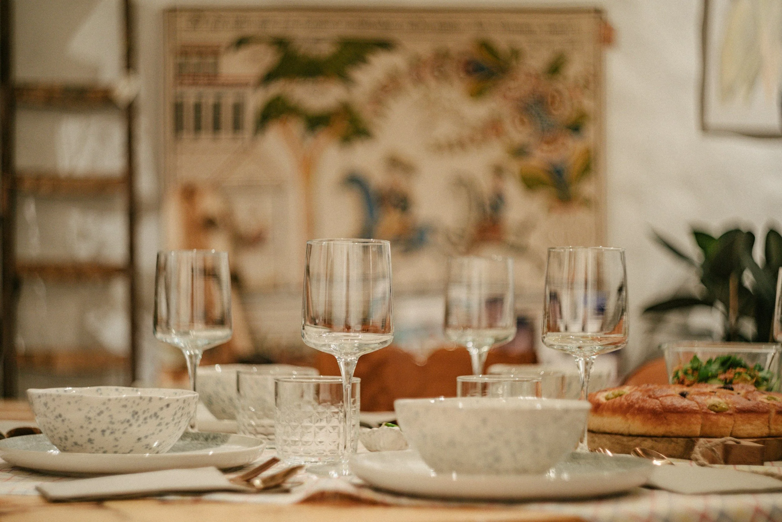 A dining table set for a meal with stemmed wine glasses, a speckled bowl, and a pizza with toppings like olives and tomatoes. In the background, abstract art hangs on the wall.