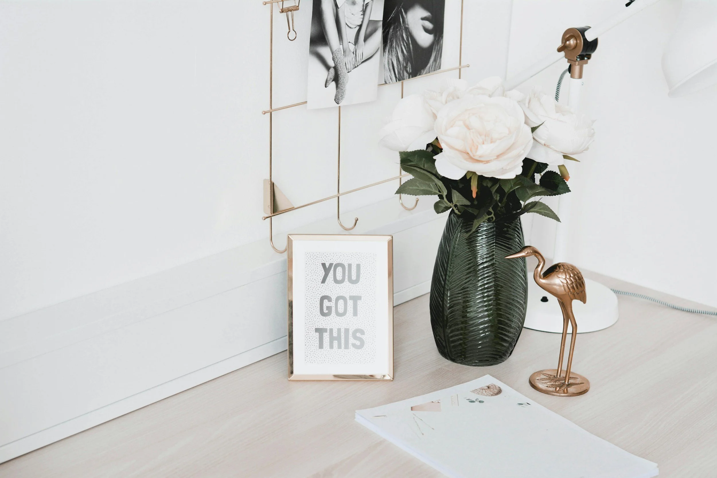 Decorative desk arrangement with black and white art photos, framed quote reading 'You got this,' a dark green vase with white floral artificial flowers, a small bronze flamingo figurine, and a magazine on a light wood surface.