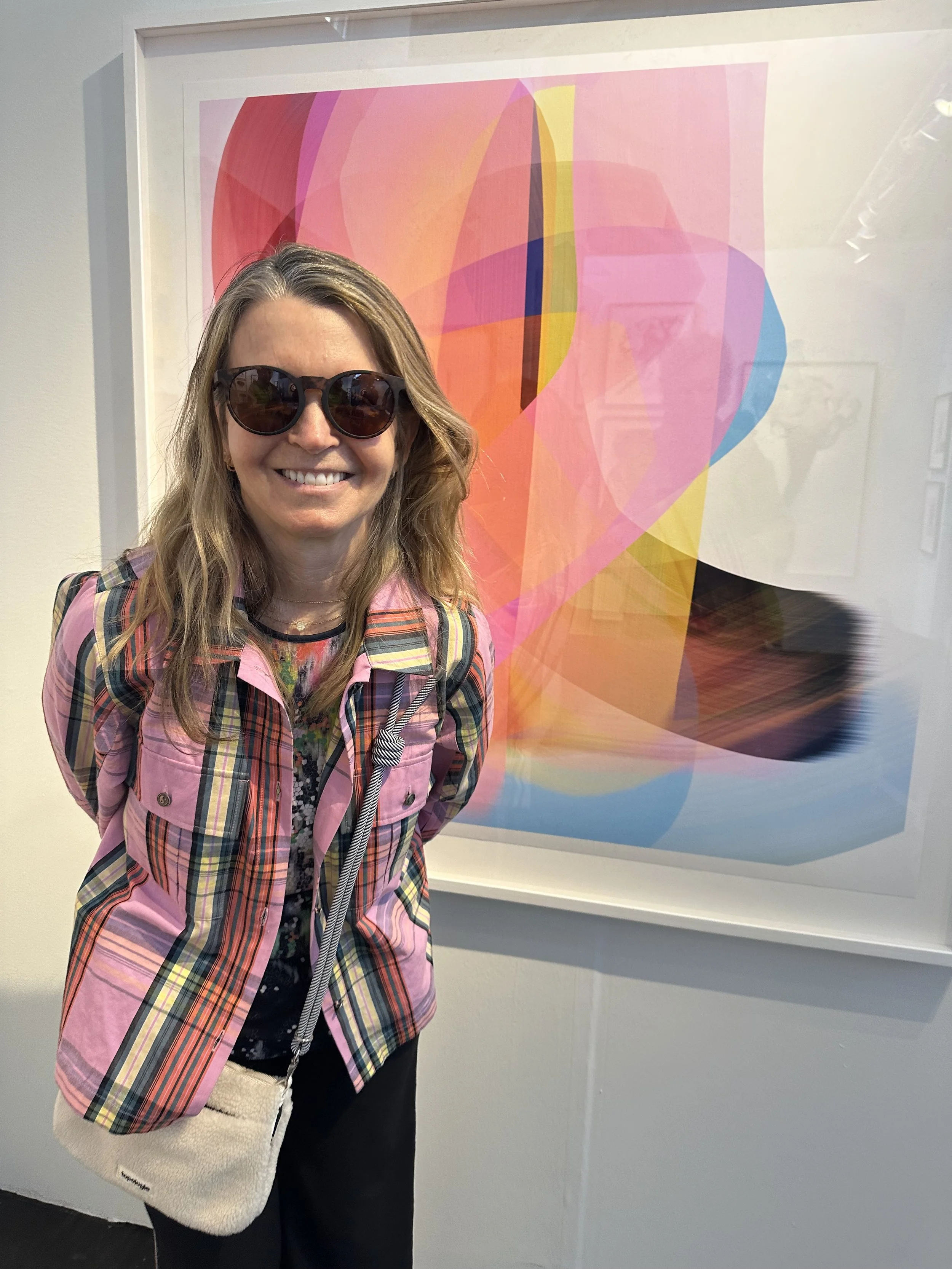 A woman with long, wavy blond hair wearing sunglasses, a pink plaid shirt, a floral top, and carrying a beige fuzzy bag, standing in front of an abstract colorful art piece.