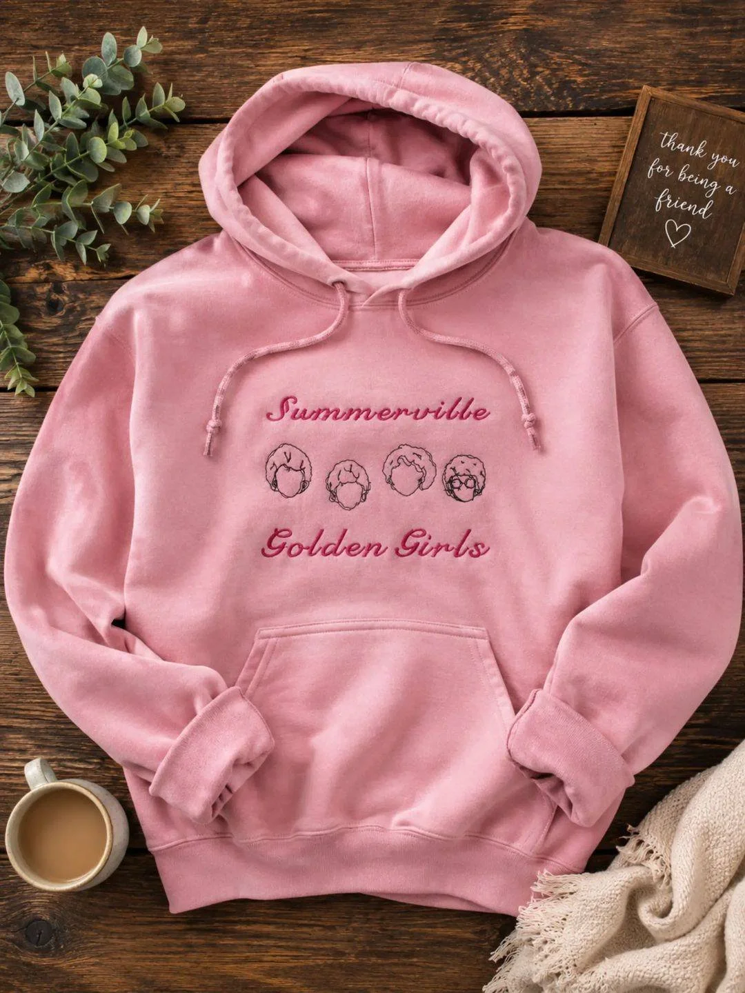 Pink hoodie with embroidered text and illustrations of four women's hairstyles. The hoodie is laid on a wooden surface with a small plant, a cup of coffee, a cream-colored scarf, and a small wooden sign that reads 'thank you for being a friend'.