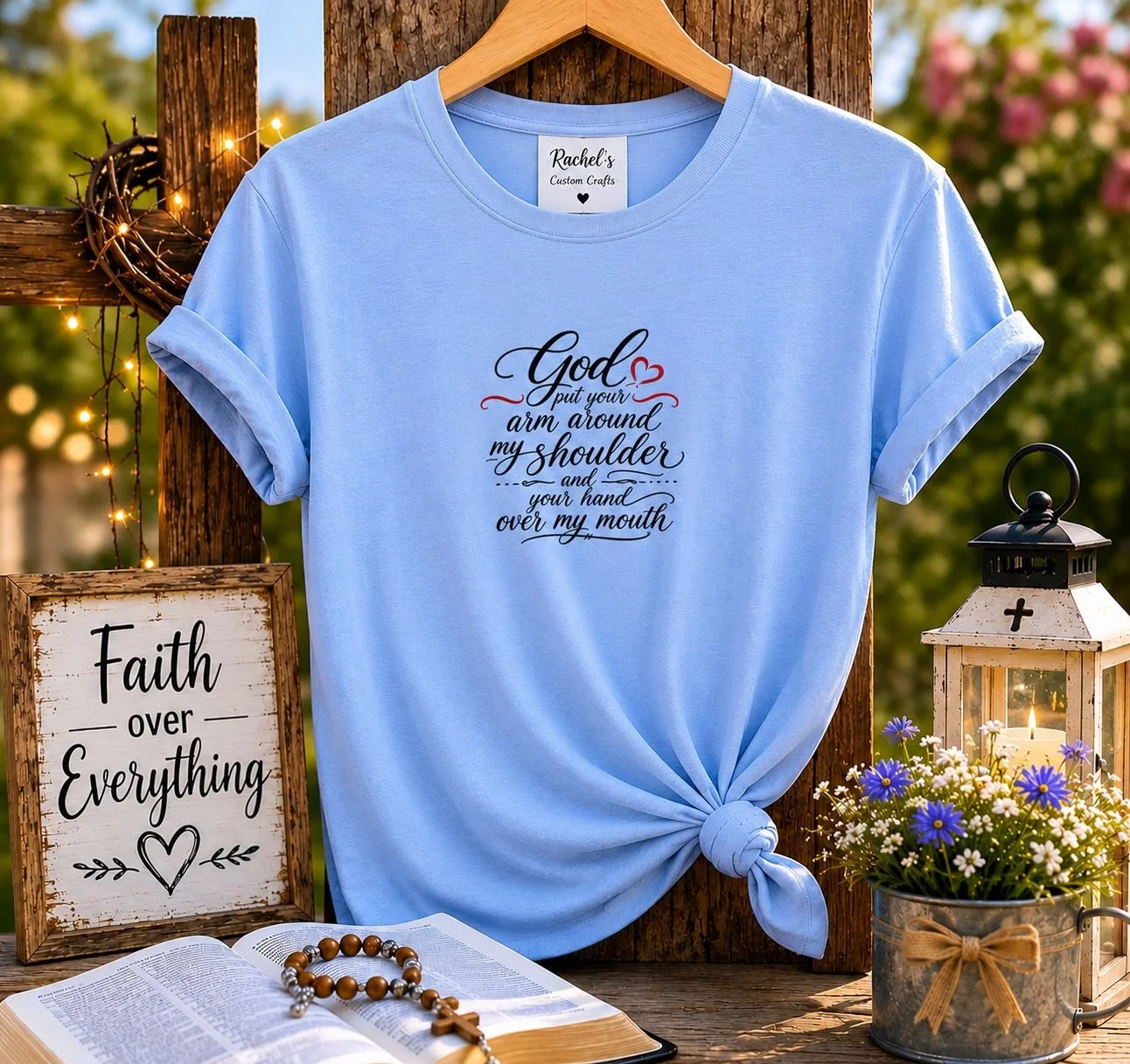 A light blue T-shirt with a religious quote hanging on a wooden post, surrounded by a lit lantern, flowers, and a sign that reads 'Faith over Everything,' in an outdoor setting.