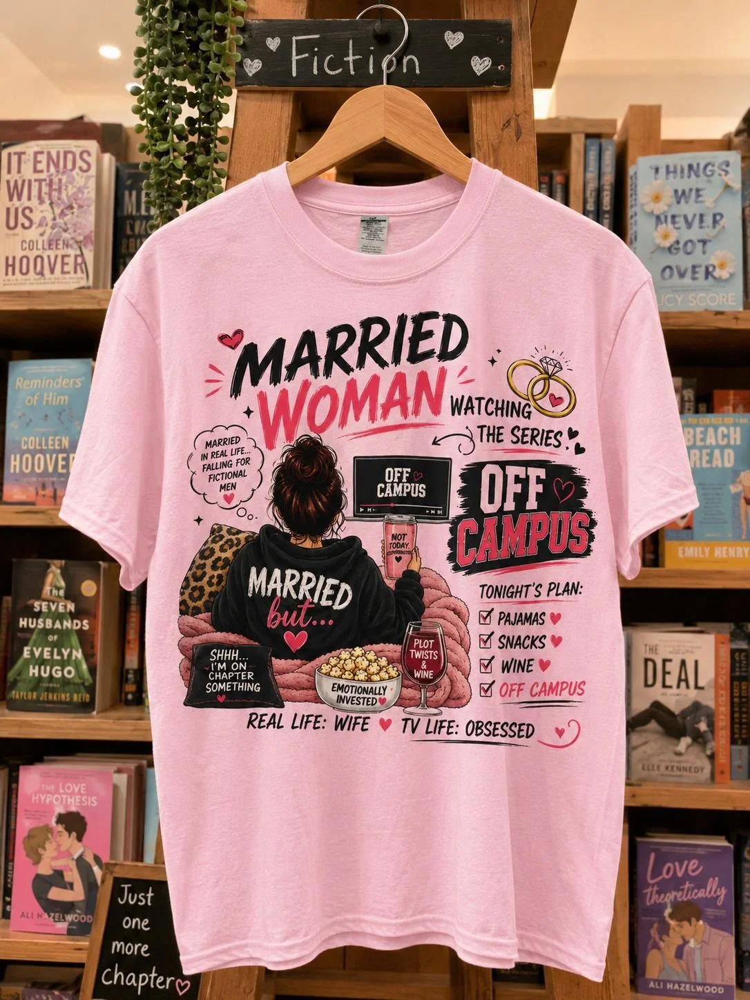 Pink T-shirt hanging on a wooden display with a sign labeled 'Fiction' at the top. The T-shirt features a cartoon woman in pajamas, holding a phone, with text and graphics about married life, watching a series, and off campus activities, with phrases like 'Married but...,' 'Tonight's plan: pajamas, snacks, wine, off campus,' and 'TV life: obsessed. Time for popcorn and wine'.