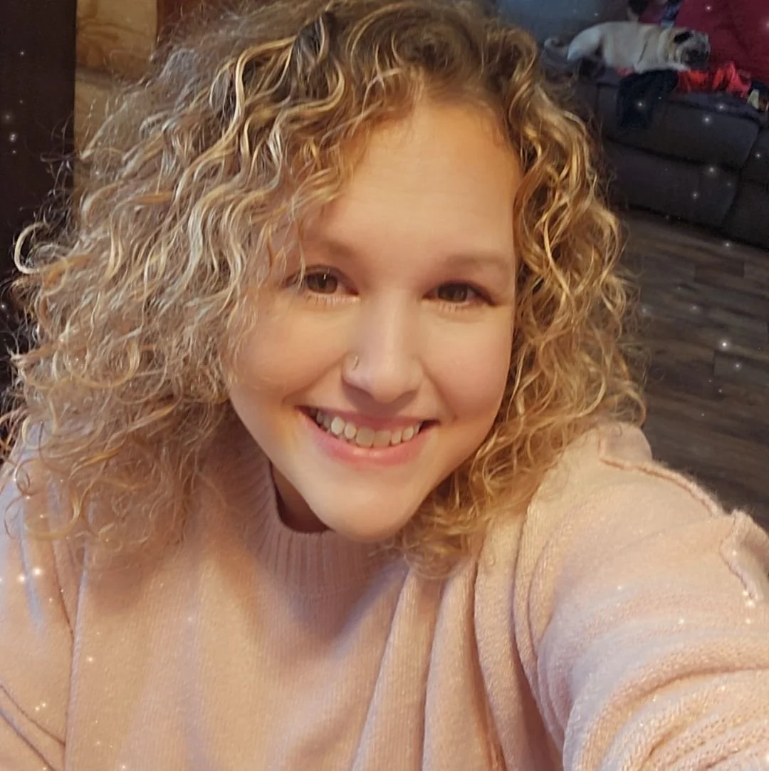 A young woman with curly blonde hair smiling at the camera, wearing a light pink sweater, in a living room with a dog lying on a couch in the background.