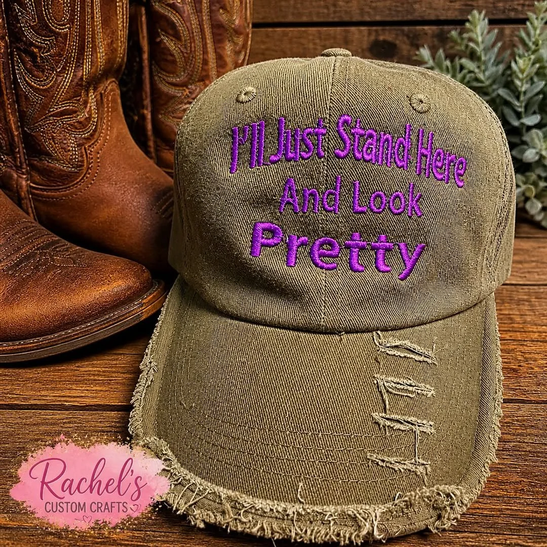 A khaki baseball cap with purple embroidered text that reads, 'I'll Just Stand Here And Look Pretty,' placed on a wooden surface. To the left, part of a brown cowboy boot is visible, and to the right, there are some green plants. A pink logo at the b