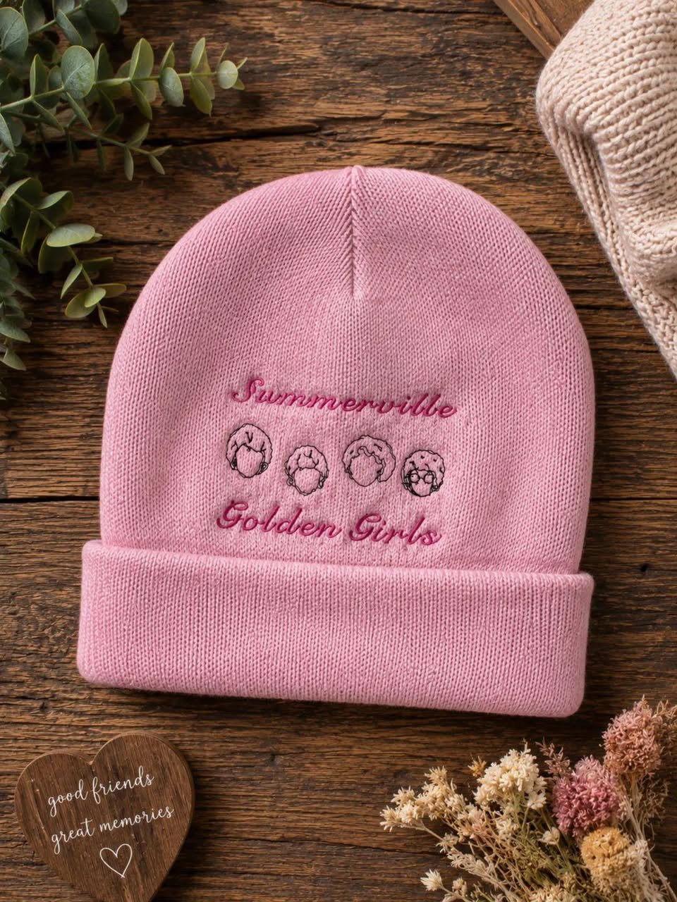 A pink knit beanie with embroidered text and illustrations of four women with different hairstyles. The text reads 'Summervibe Golden Girls'. The beanie is on a wooden surface alongside dried flowers and a small wooden heart with the words 'good frie