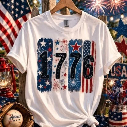 White T-shirt with a patriotic design featuring the date 1776, decorated with stars, fireworks, and American flag patterns, hung against a backdrop of patriotic decorations and American flags.