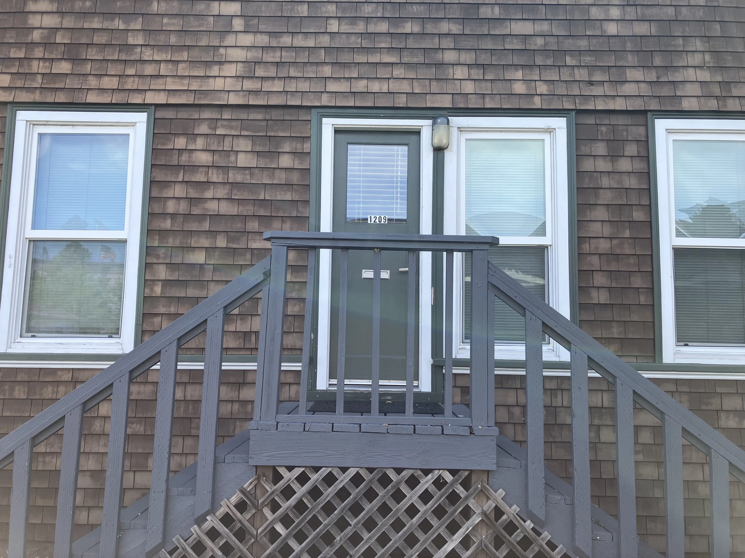 A house front with brown shingle siding, three windows with white trim, a small door with the number 1209, and a gray wooden staircase leading up to the door.