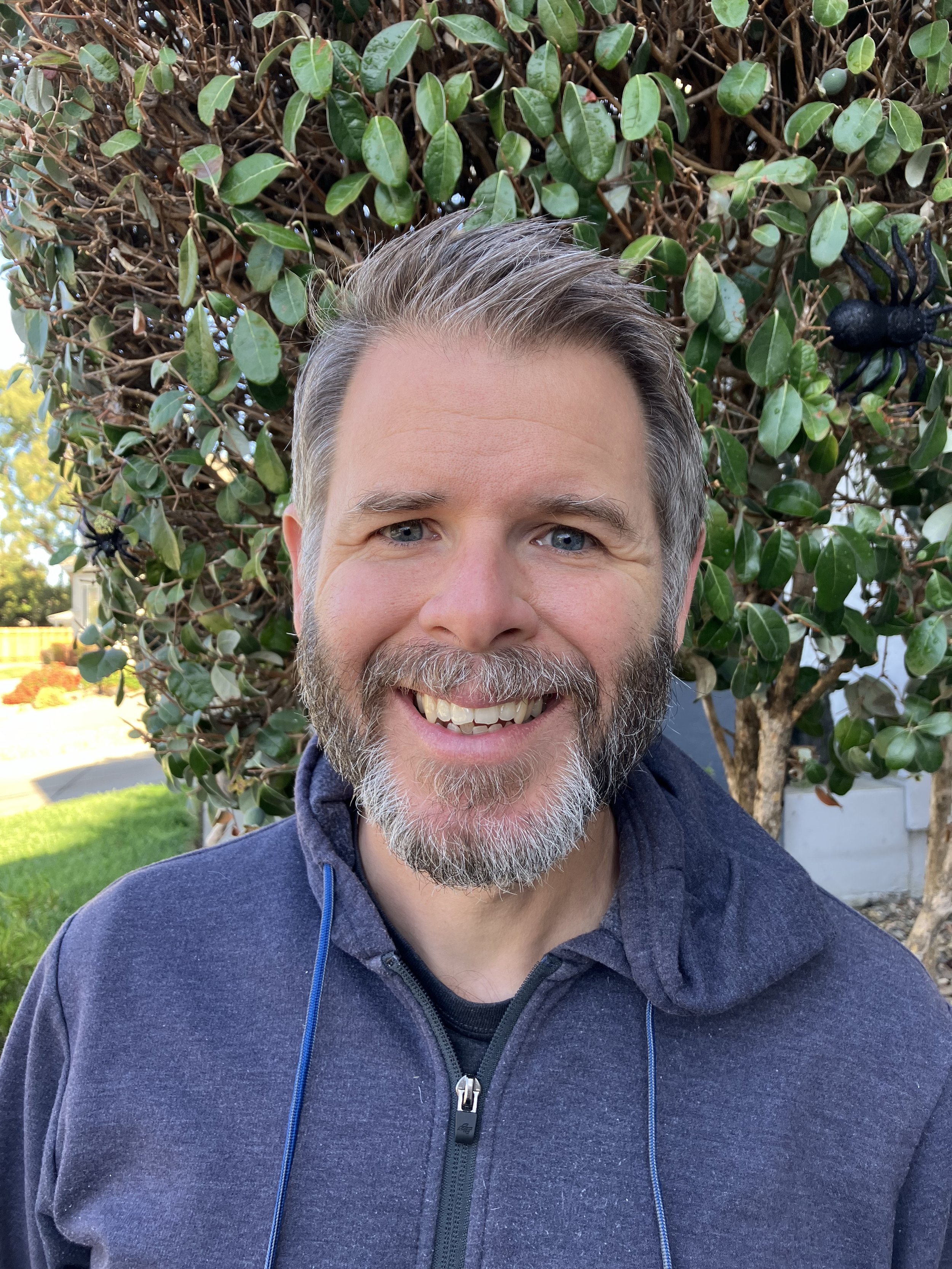 A man with gray hair and beard smiling outdoors in front of a bush with green leaves and a black plastic toy spider. He is wearing a navy blue zip-up jacket.