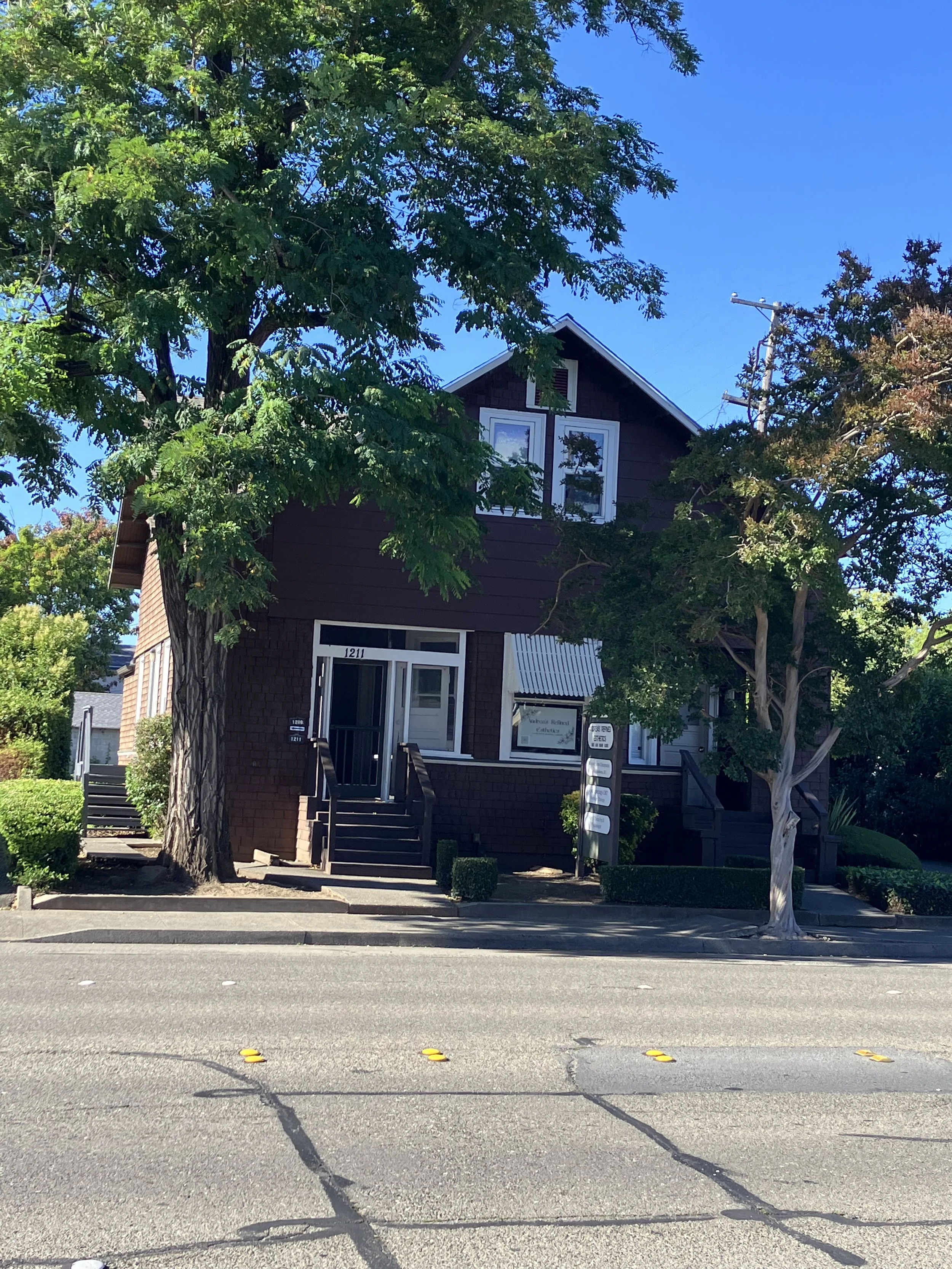 A two-story house with dark exterior siding, white window frames, and a small front porch with stairs. There are tall trees with green foliage partially obscuring the house and a clear blue sky. The street in front has yellow parking blocks and a sidewalk with some small bushes.