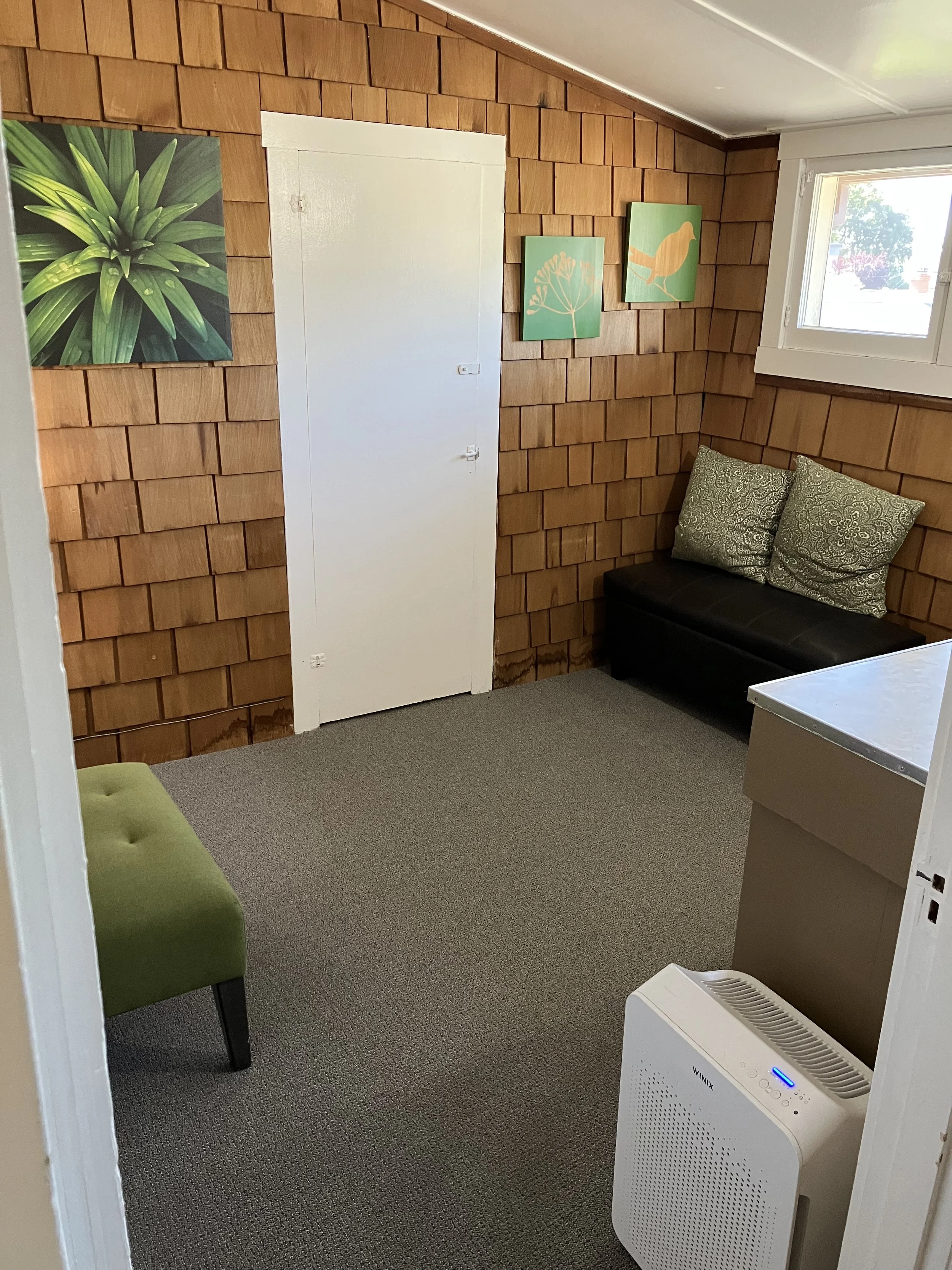 A small cozy room with wooden shingle walls. There is a black bench with two decorative pillows, a window letting in natural light, a small green cushion on a piece of furniture, and a white portable air purifier in the corner. Three pieces of artwork featuring botanical and bird illustrations hang on the wall.