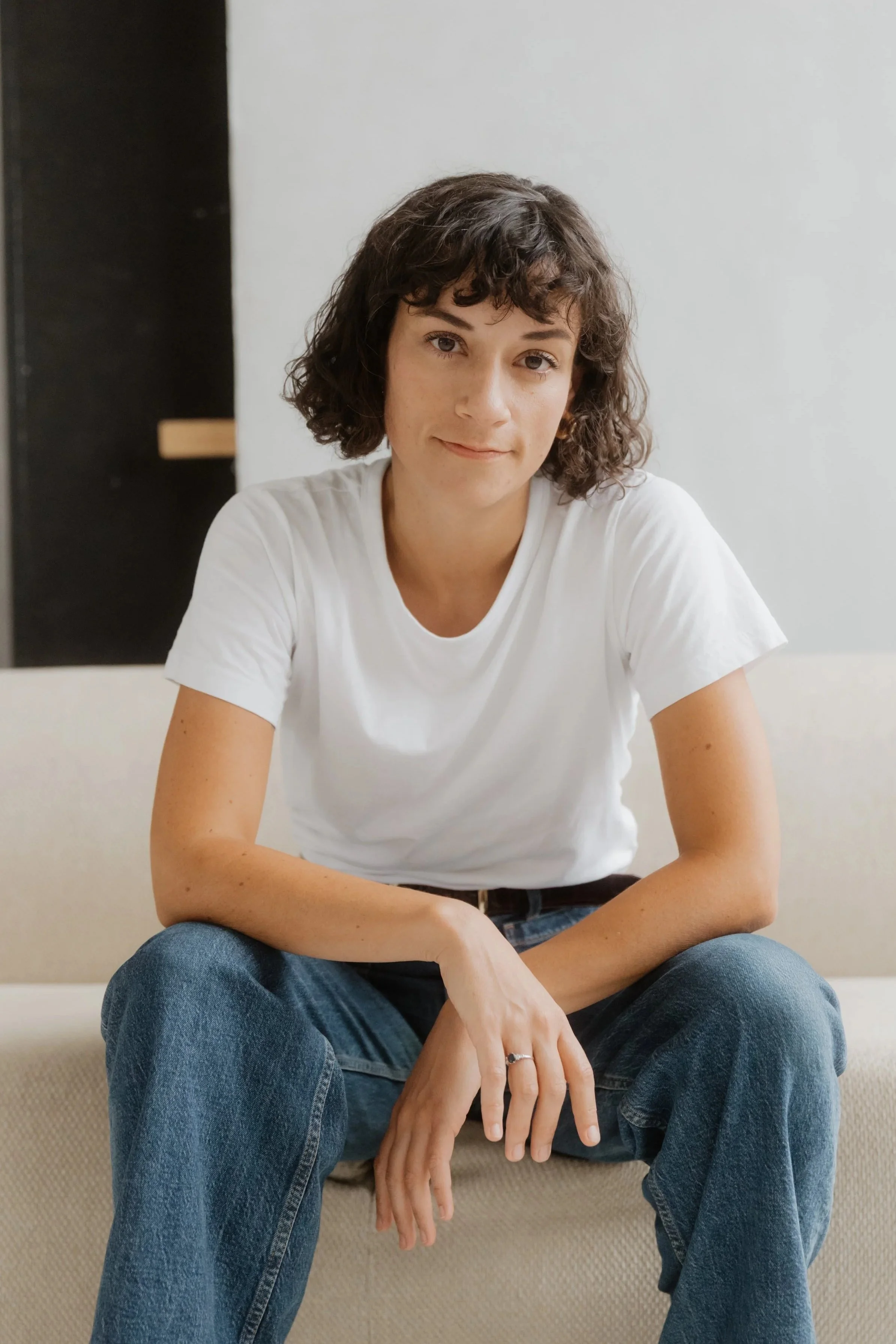 Junge Frau mit dunkelbraunen, lockigen Haaren, trägt ein weißes T-Shirt und sitzt auf einem Sofa, schaut in die Kamera, lächelt leicht, in einem hellen Raum.