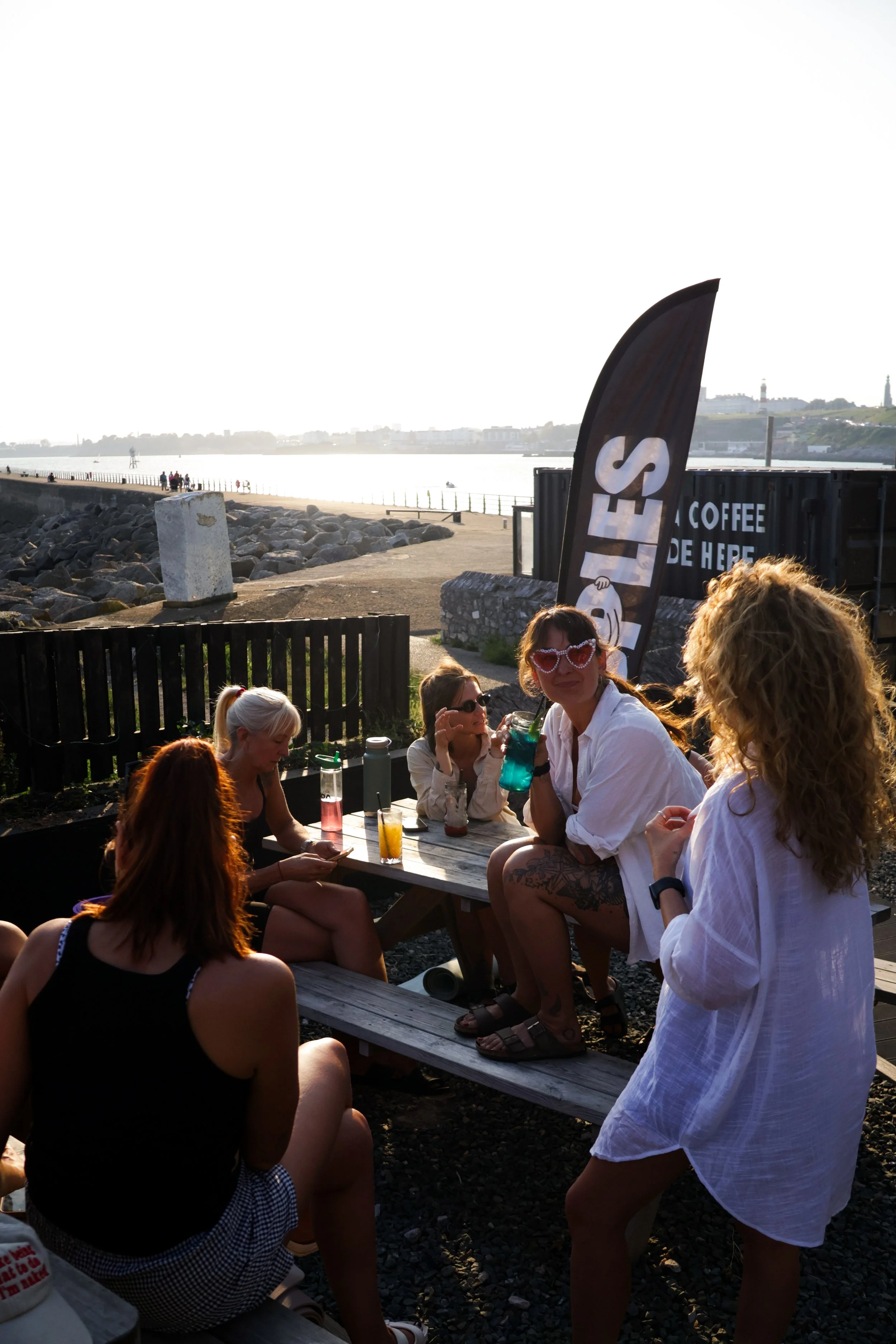 Group of people sitting and standing around a picnic table outdoors near a waterfront during sunset, with some holding drinks and wearing casual summer clothes.