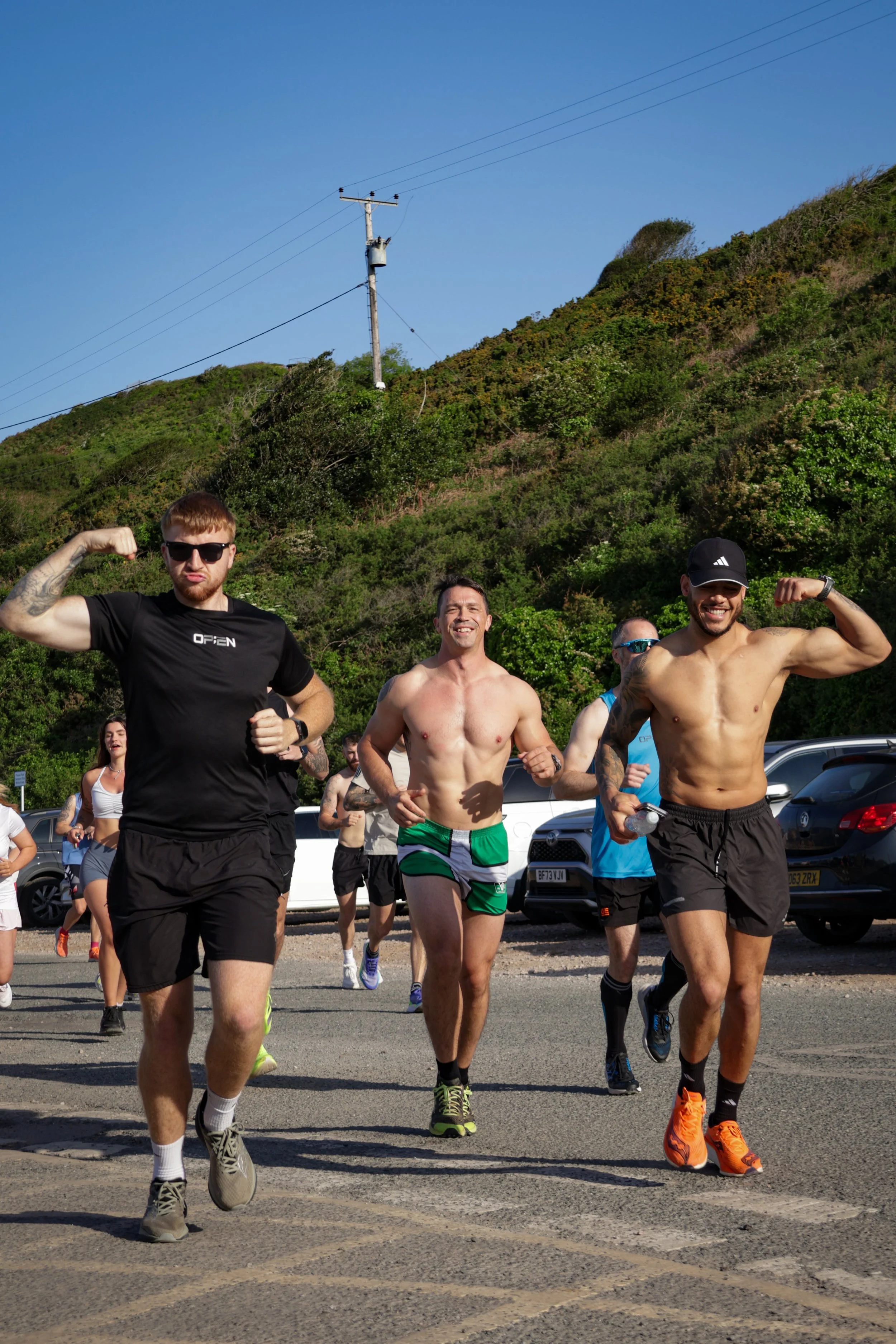 A group of men and women participating in an outdoor running event on a sunny day, with some men in the foreground showing muscular poses. The background features lush green hills, parked cars, and a utility pole against a clear blue sky.