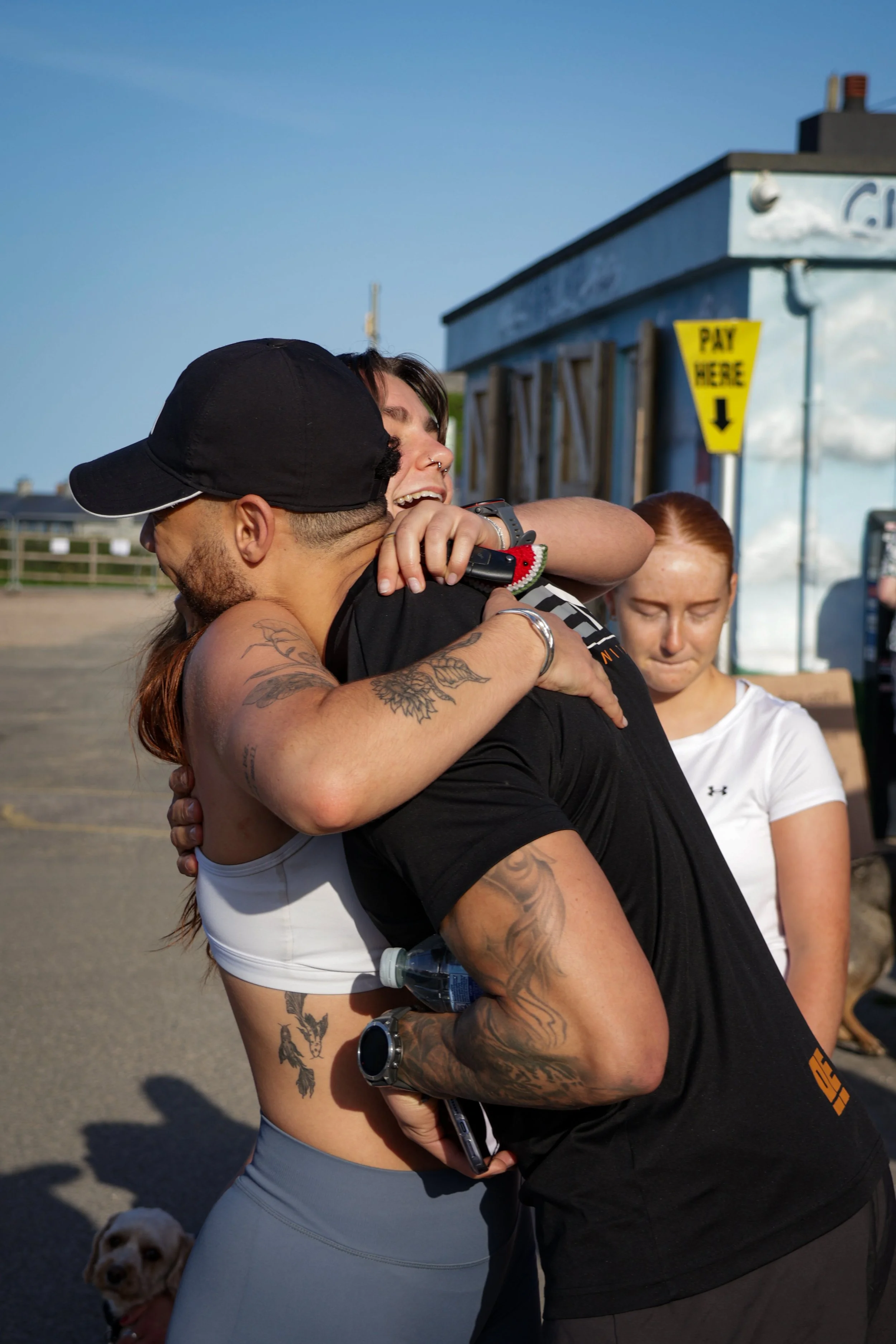 Two women hugging outdoors on a sunny day, with a small dog in the background, and a blue building with a 'Pay Here' sign in the distance.