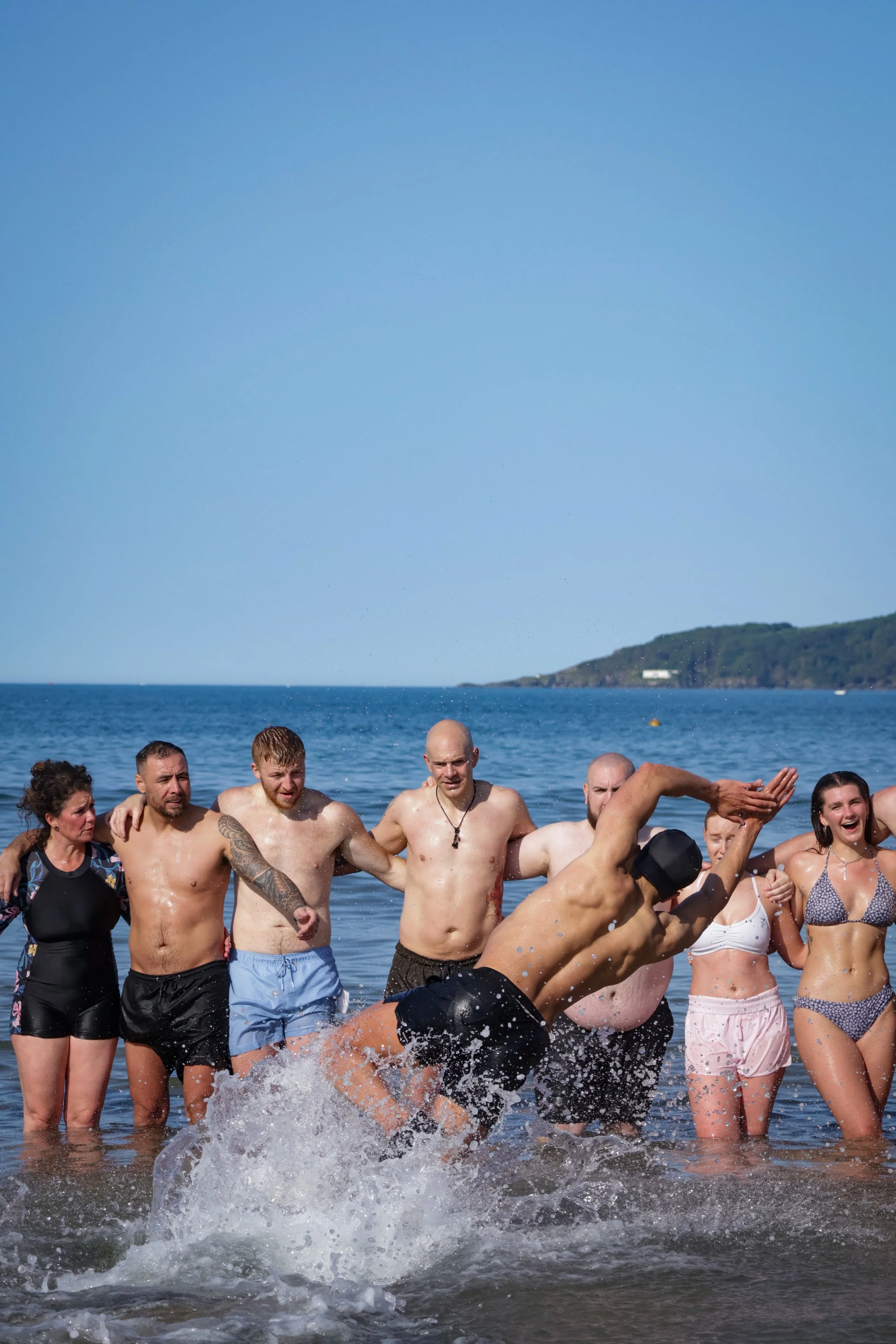 Group of people swimming and playing in the ocean near the shoreline on a sunny day, with clear blue sky and distant land in the background.