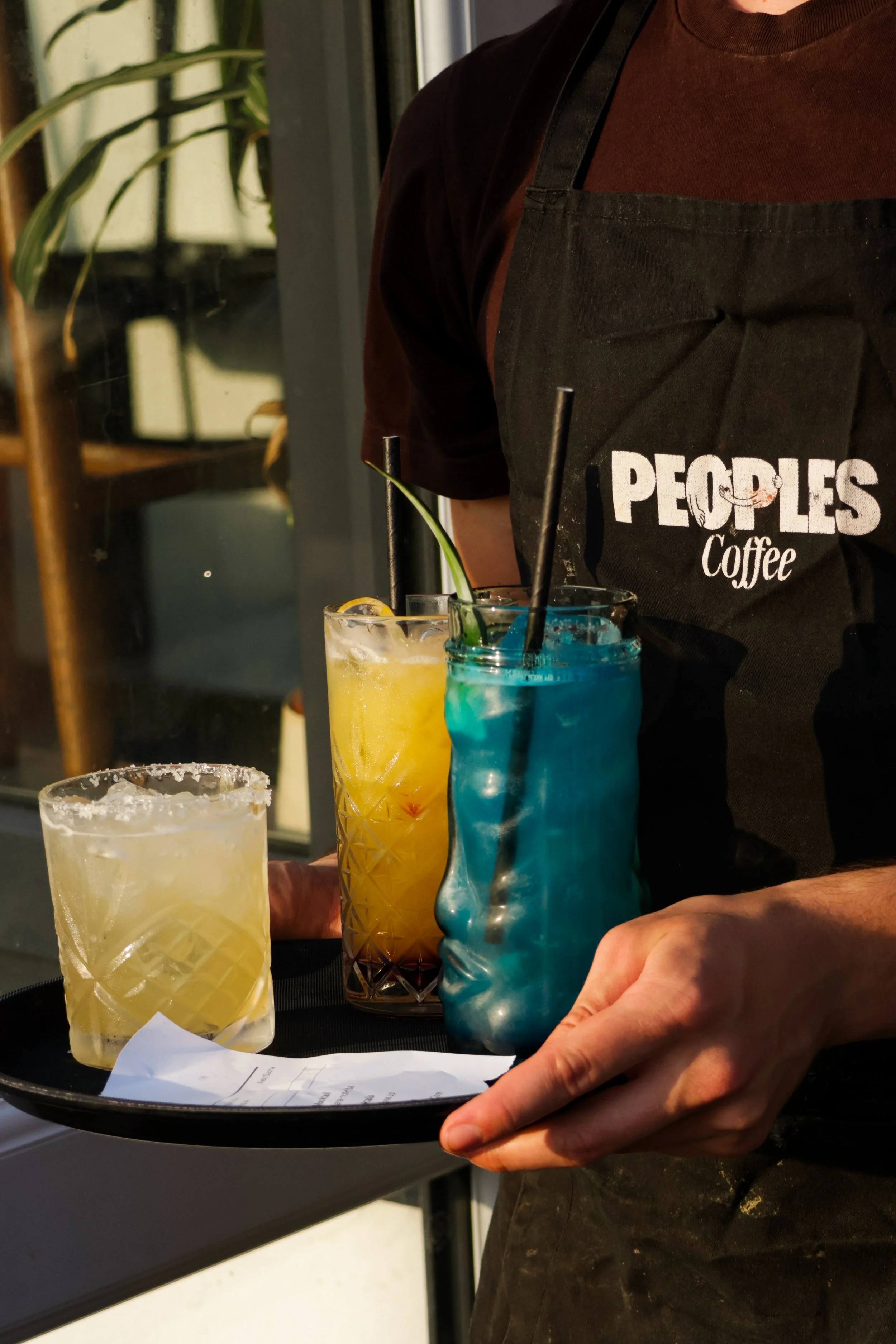 Person wearing a black apron with the words 'PEOPLES Coffee' in white, holding a tray with three colorful drinks, including a yellow cocktail with a lemon garnish and a blue drink in a carved glass, inside a coffee shop.