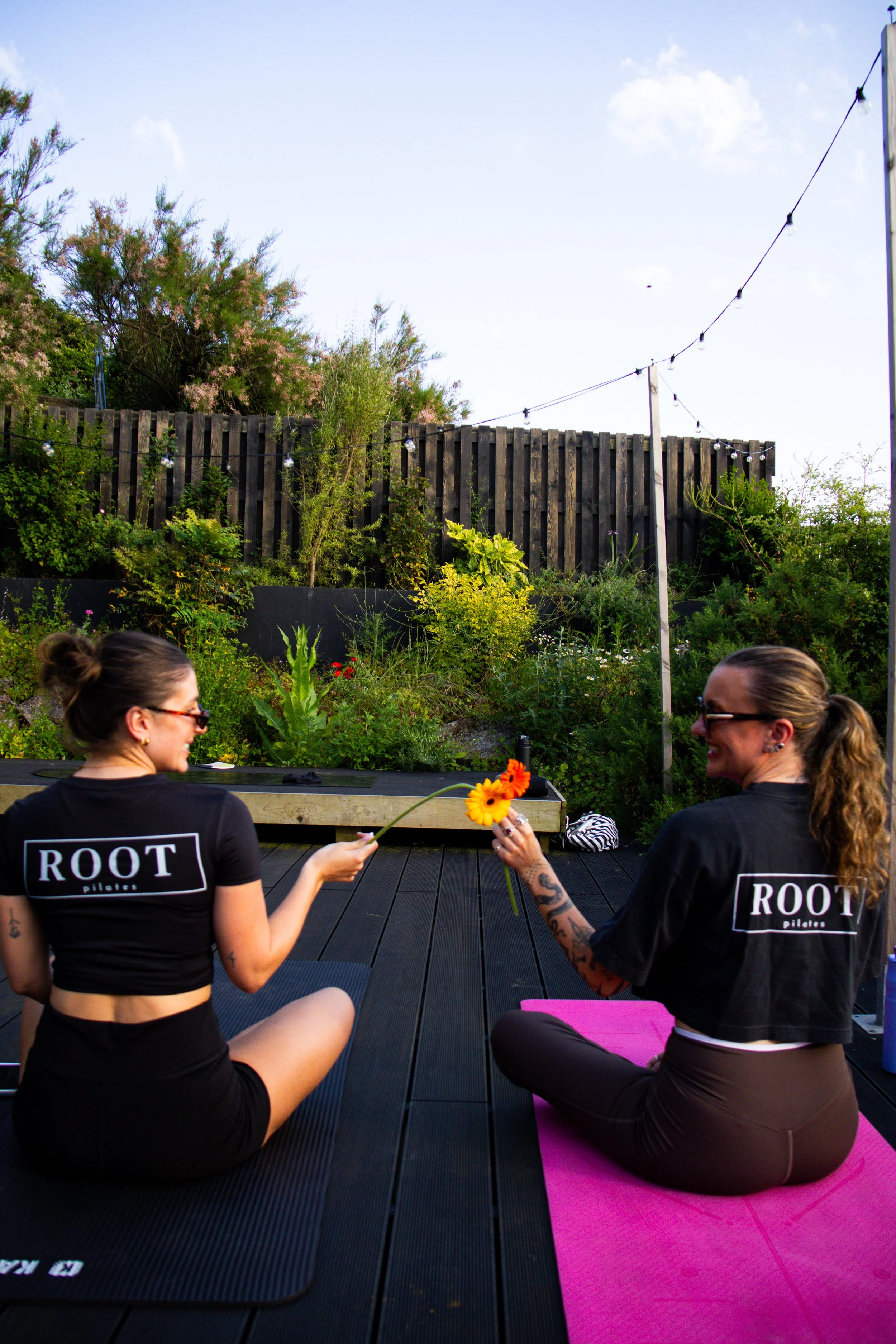 Two women practicing yoga outdoors on a wooden deck, sitting cross-legged on yoga mats, holding flowers, with a decorative fence, trees, and string lights in the background.