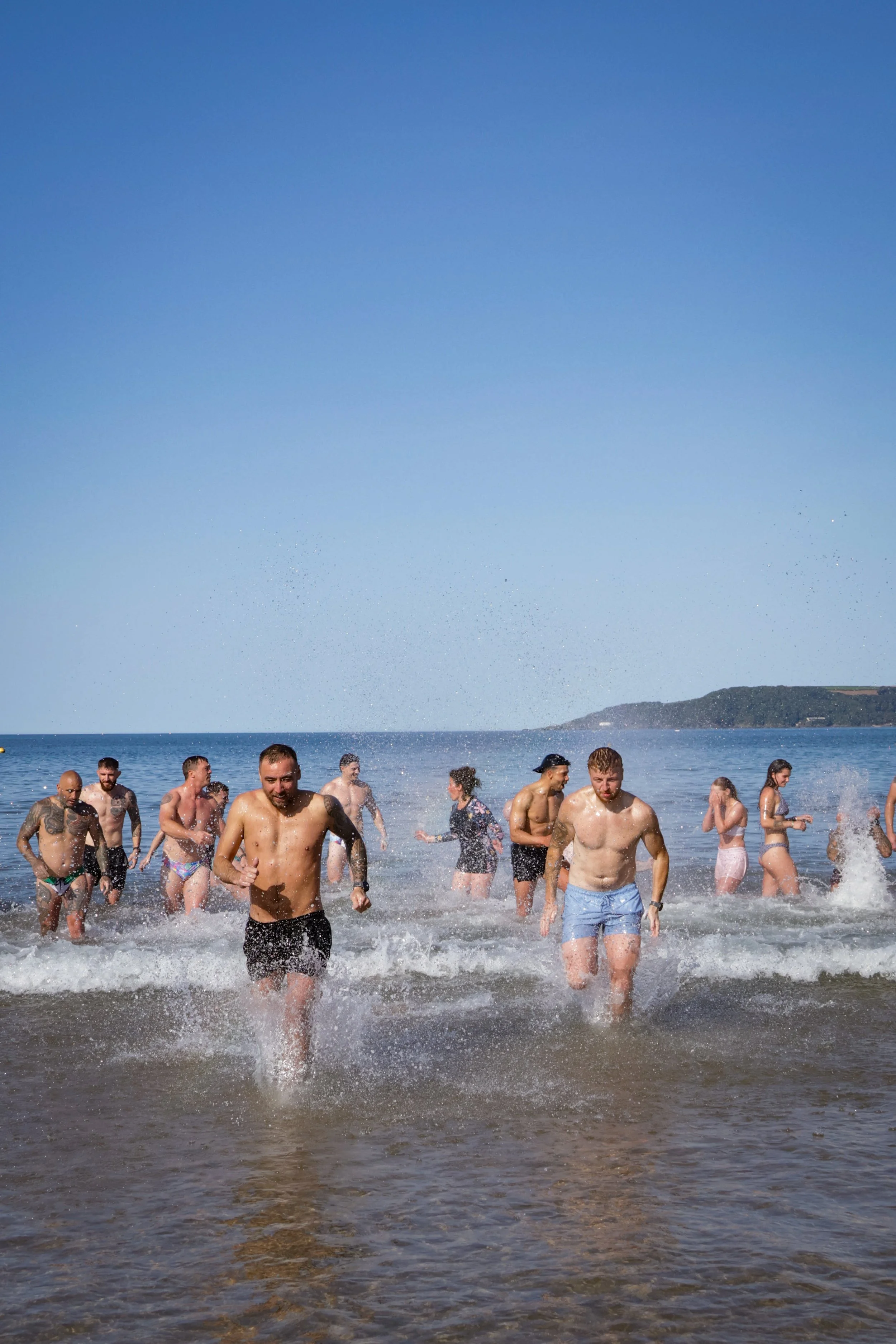 People are swimming and playing in the ocean near a beach with a hill in the background under a clear blue sky.