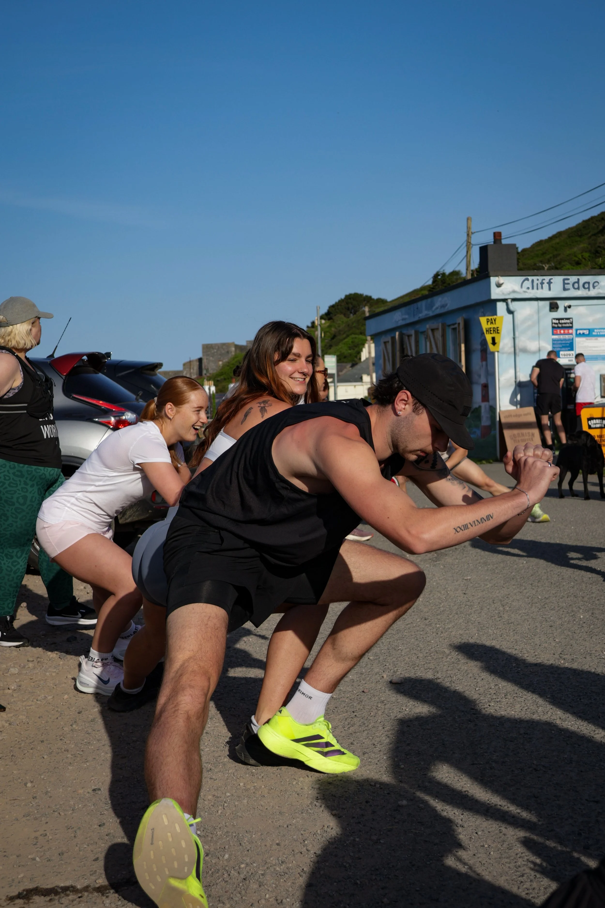 A group of people participating in an arm-wrestling contest outdoors on a sunny day. They are pulling on each other's hands while bending forward, with smiling faces and a clear blue sky in the background.