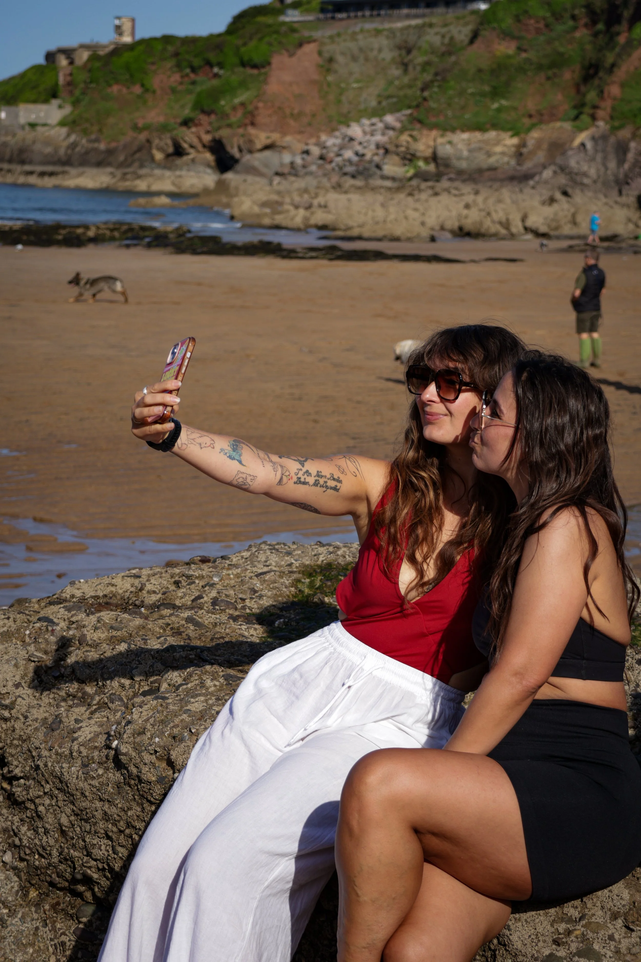 Two women sitting on rocks at the beach, taking a selfie. One woman has long curly brown hair, wearing sunglasses, a red top, and a white skirt. The other woman has long dark hair, wearing a black top. In the background, there are people walking on t