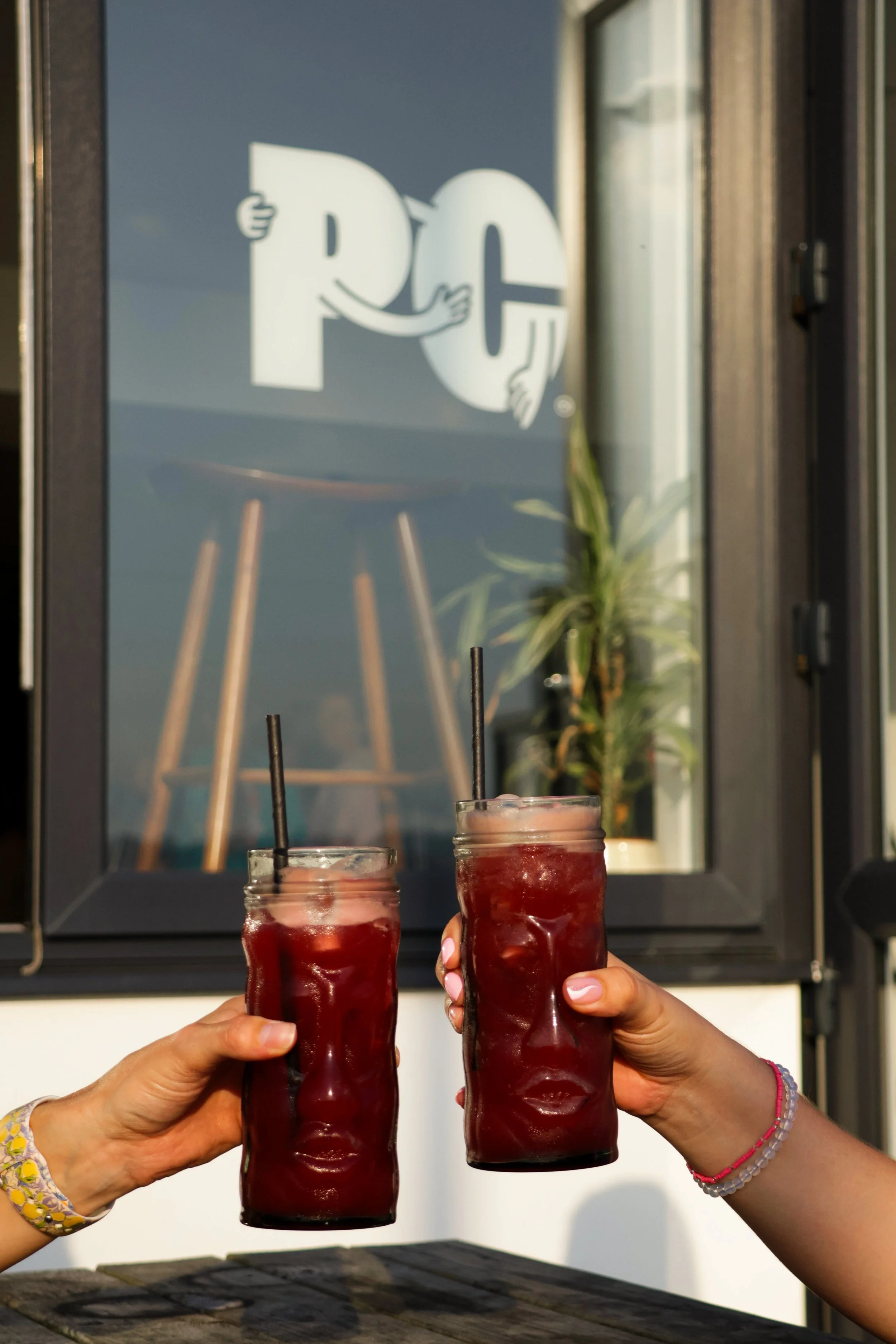 Two people clinking glasses of red drinks outdoors, with a reflective window and a plant in the background.