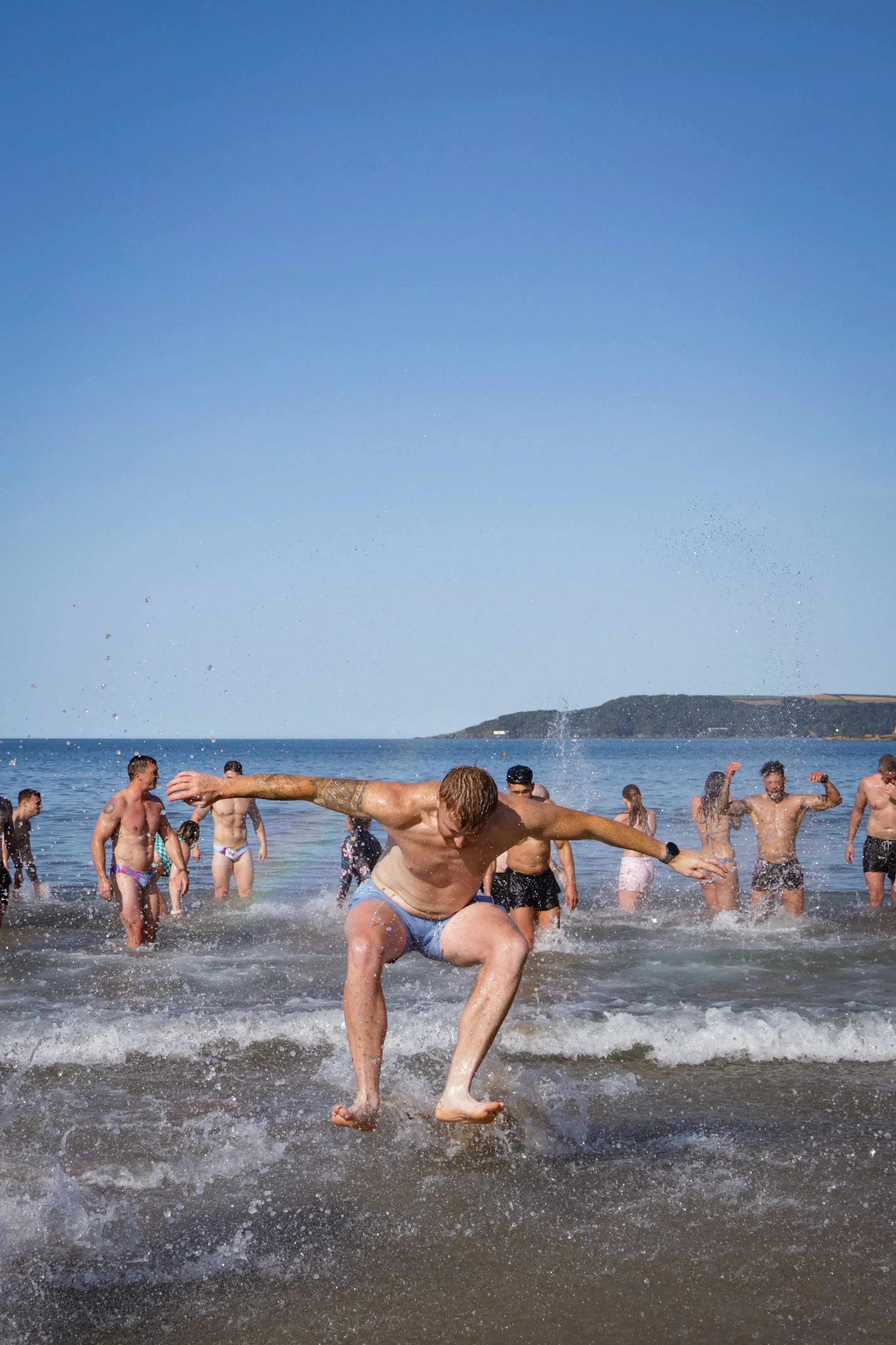 People enjoy a fun day at the beach, splashing and playing in the water on a sunny day with a clear blue sky and a distant landmass in the background.