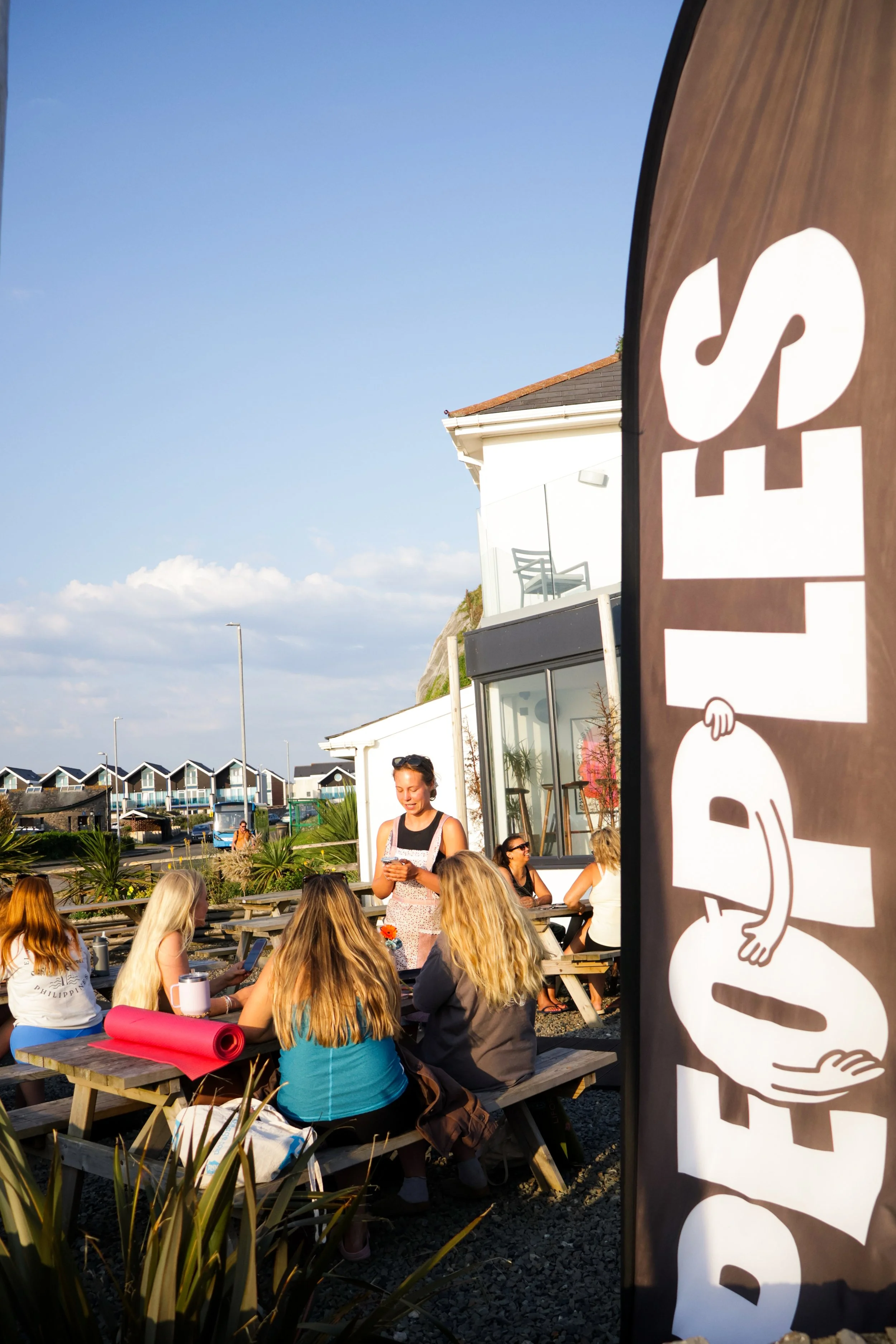 A group of people sitting at picnic tables outdoors, with a person standing and interacting with them, under a clear blue sky. There is a large sign with a stylized hand holding a cup, likely representing a coffee shop or café, partially visible on t