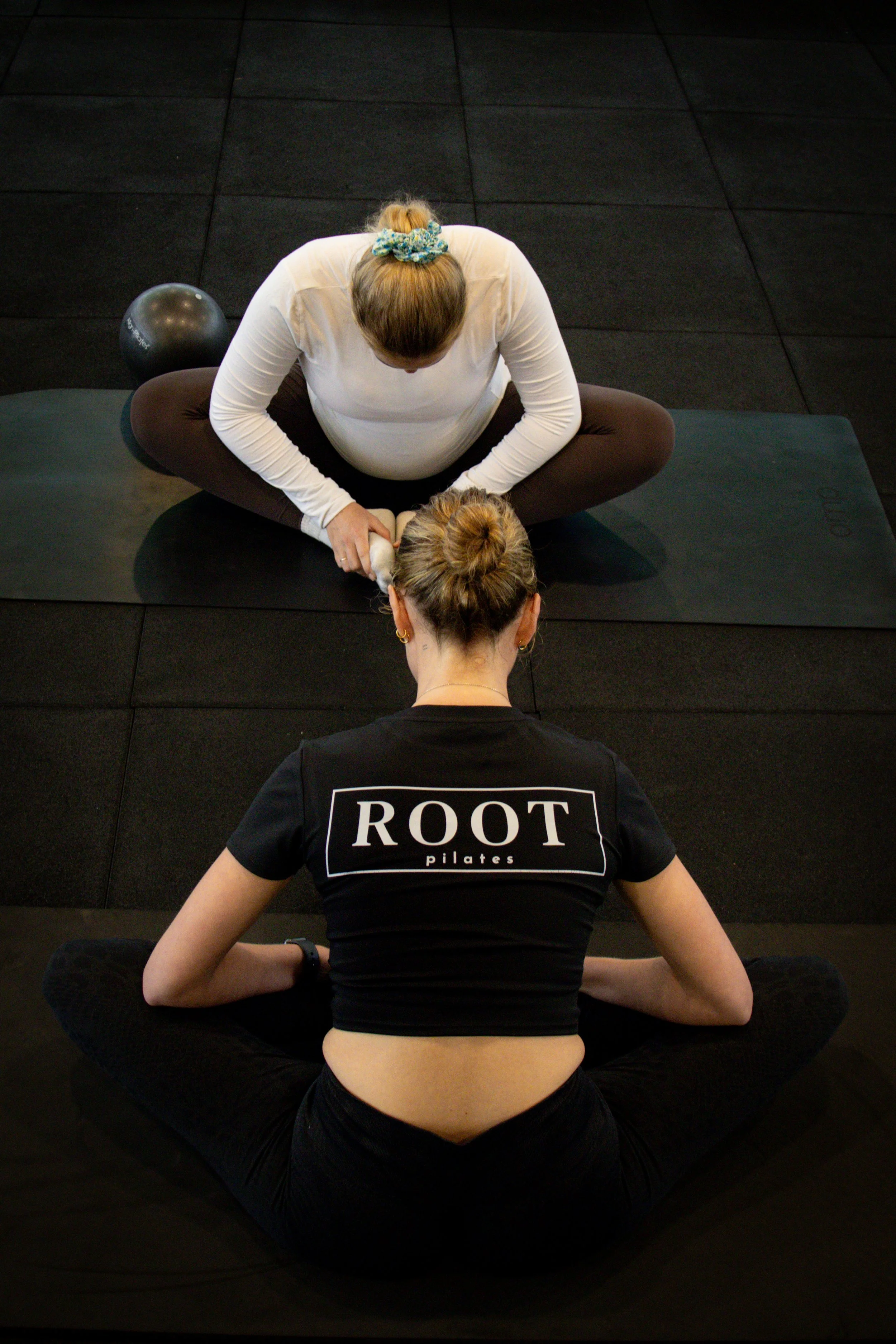 Two women practicing yoga or meditation in a gym with black mats. One woman is wearing a black shirt with 'ROOT pilates' and the other is wearing a white long-sleeve shirt. They are sitting cross-legged with hands resting on their knees, focusing dow