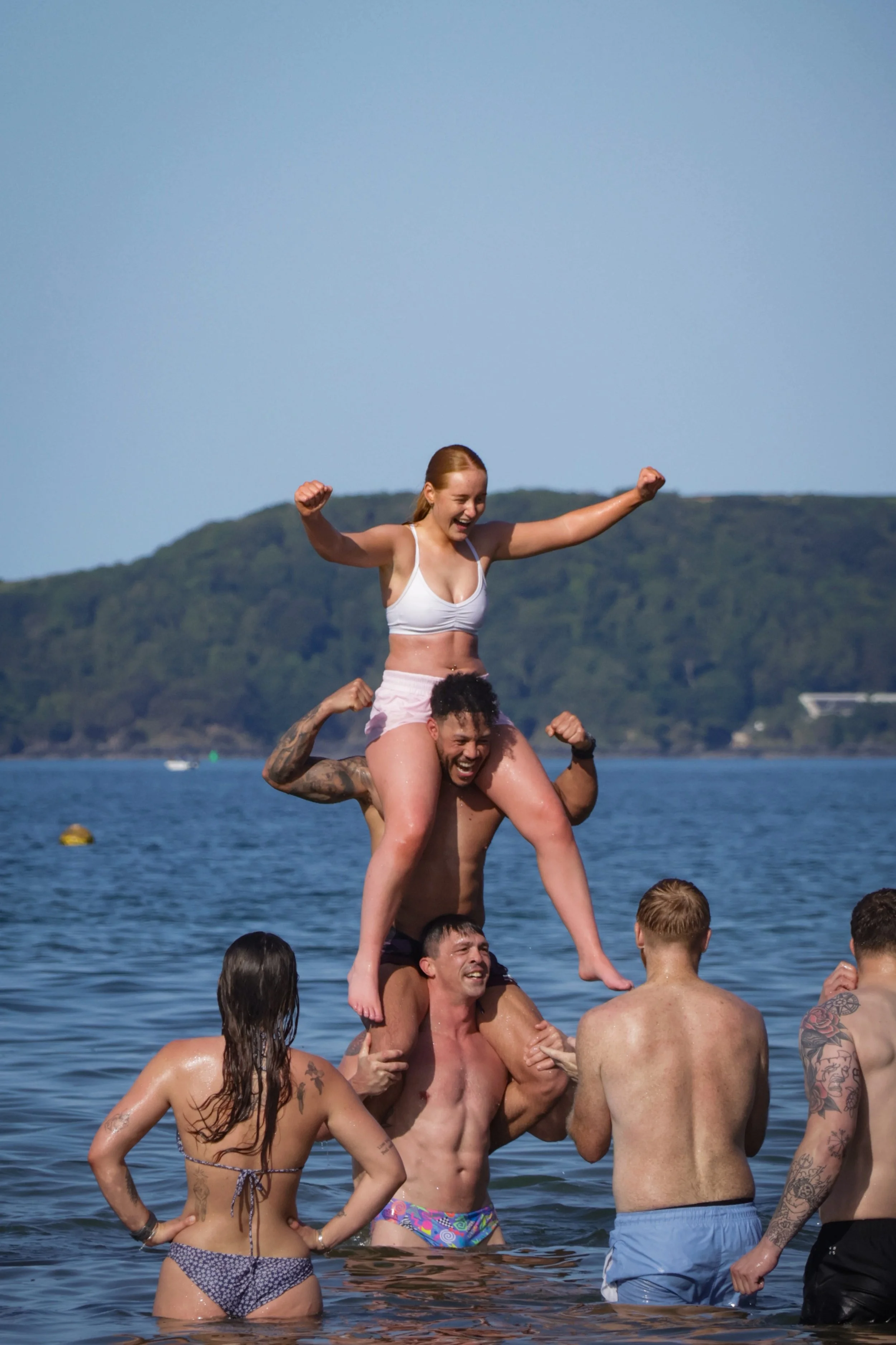 People in swimsuits celebrating and forming a human pyramid in a body of water with a hilly landscape in the background.