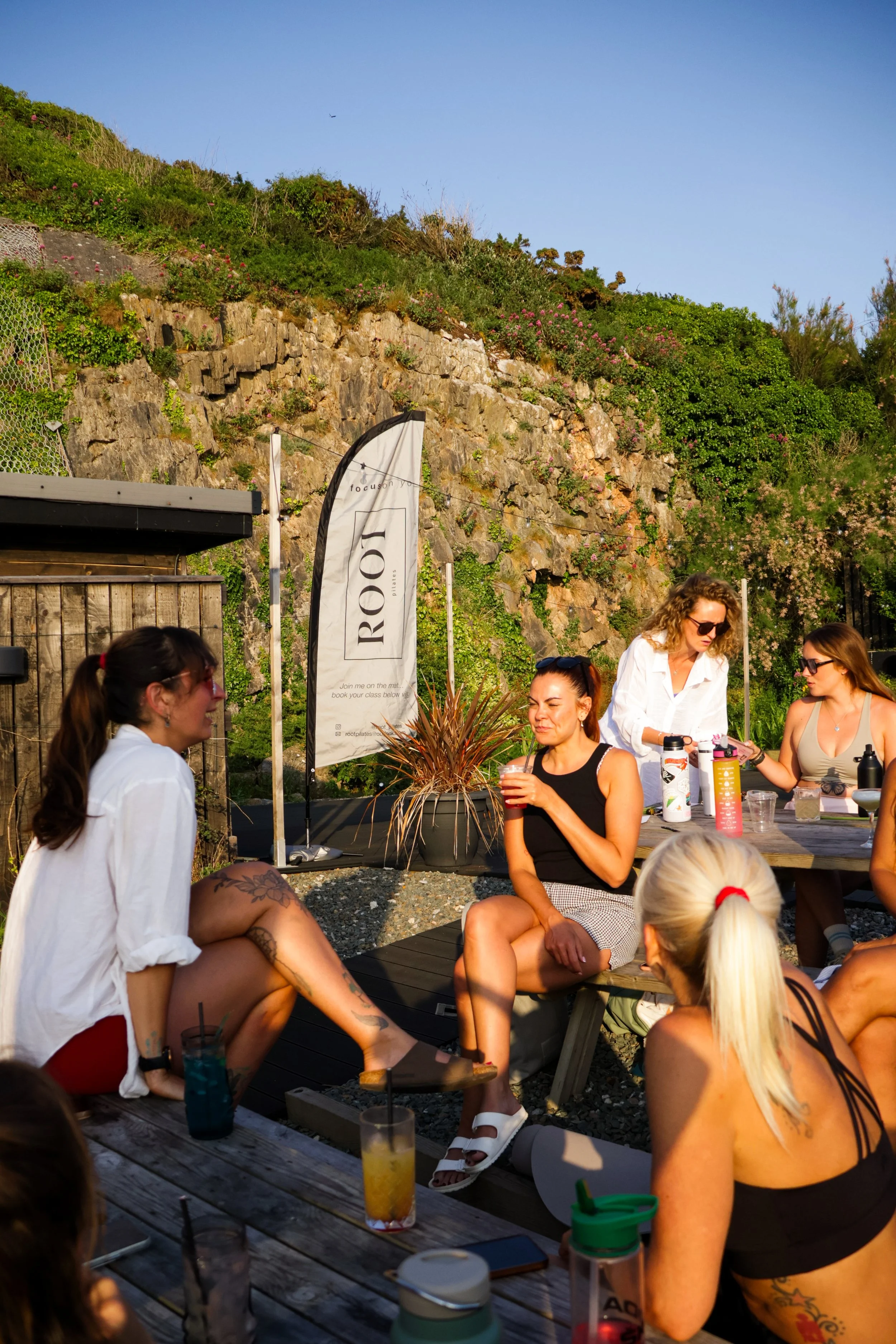 Group of women socializing outdoors at a wooden picnic table near a hillside with green bushes and rocks, sunny weather, some drinks and food on the table, and a banner with the word 'ROBE' in the background.
