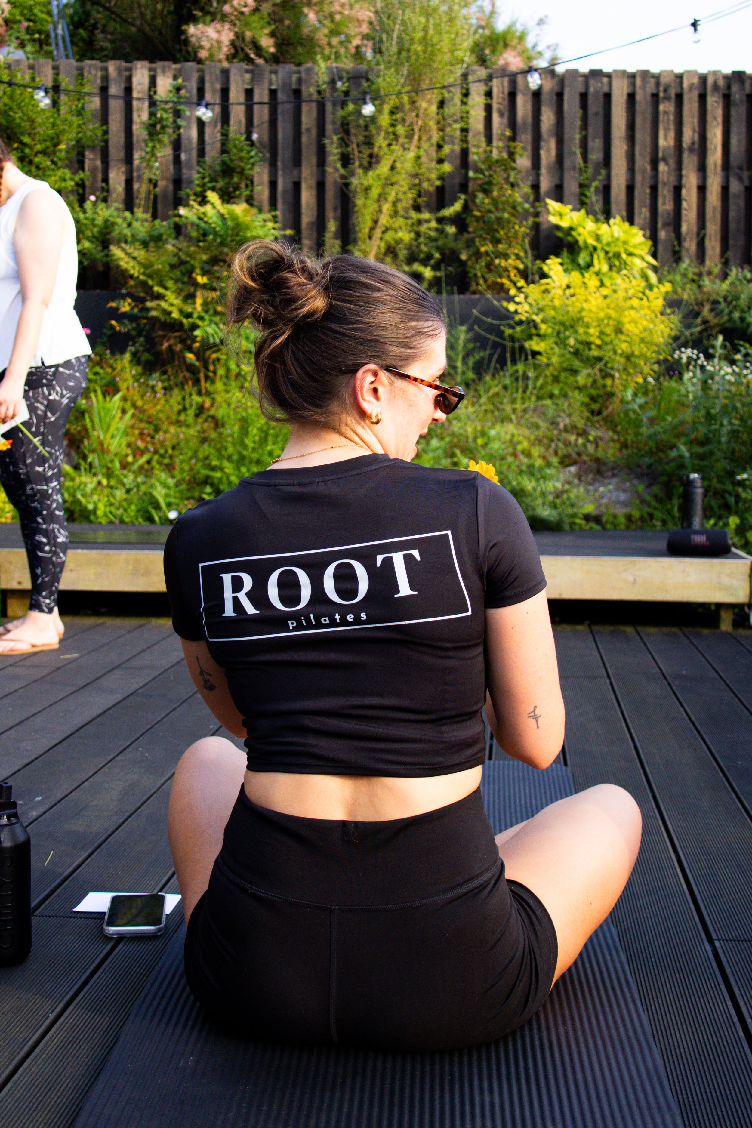 A woman with brown hair in a bun, wearing sunglasses and a black 'ROOT pilates' t-shirt, sitting on a black yoga mat outdoors, with a garden and wooden fence in the background. Another person is visible partially on the left side of the image.