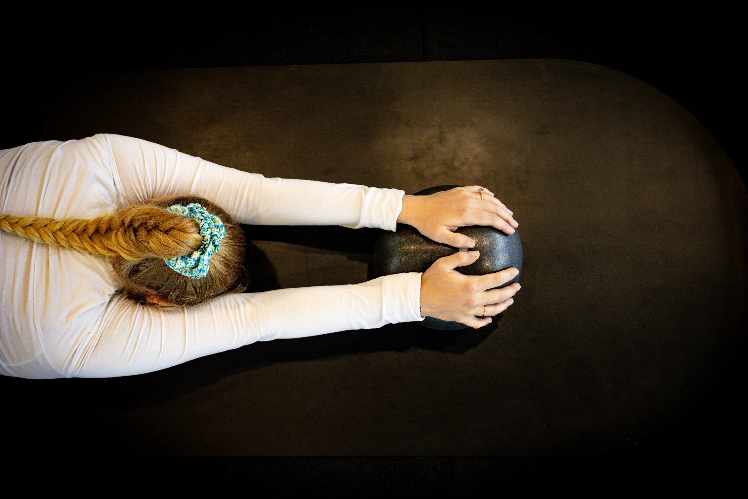 A woman lying on her side on a black therapy mat, with her head turned away, holding a black exercise ball with both hands, in a dark room.