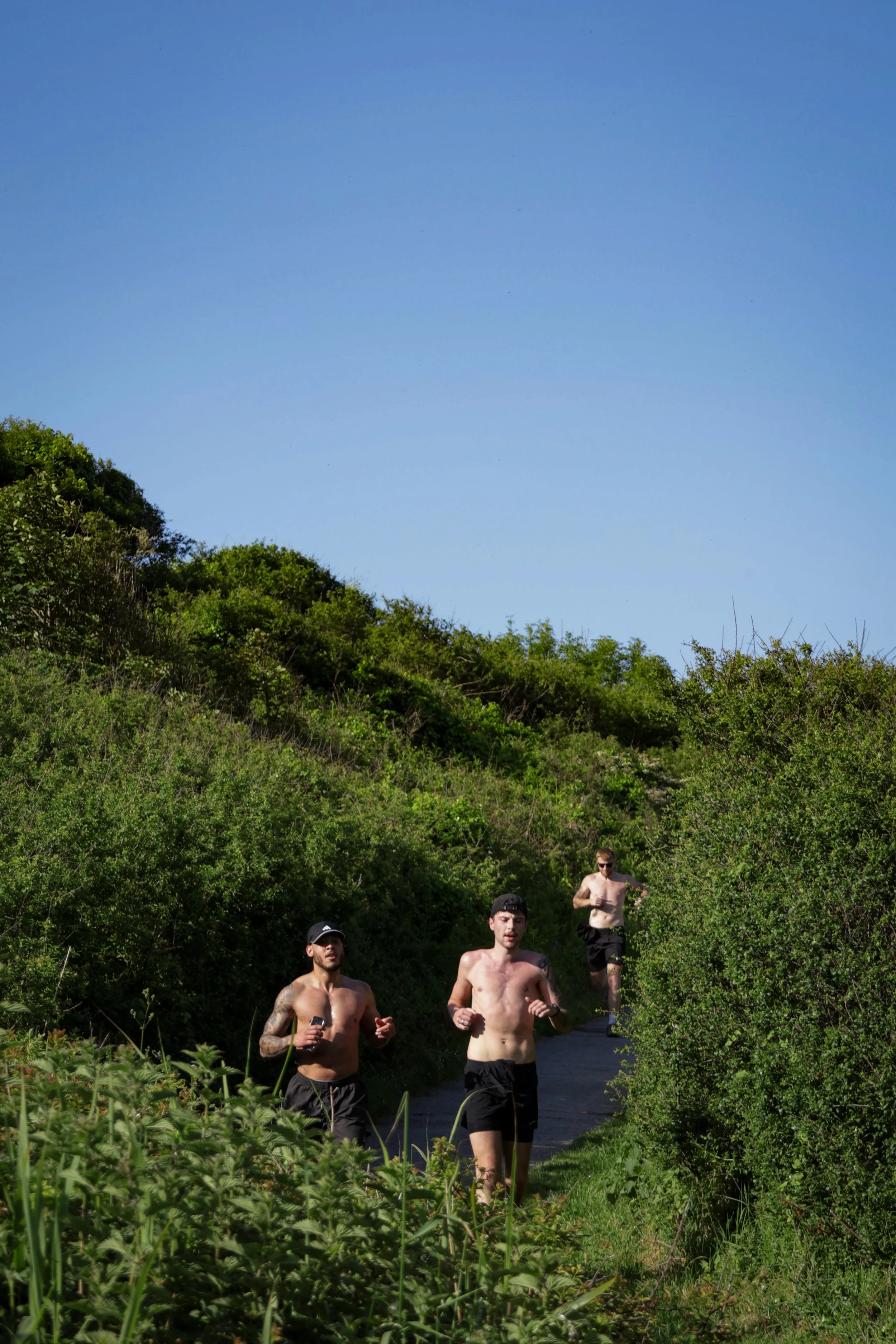 Group of four shirtless men running on a forest trail with lush green bushes and trees, under a clear blue sky.
