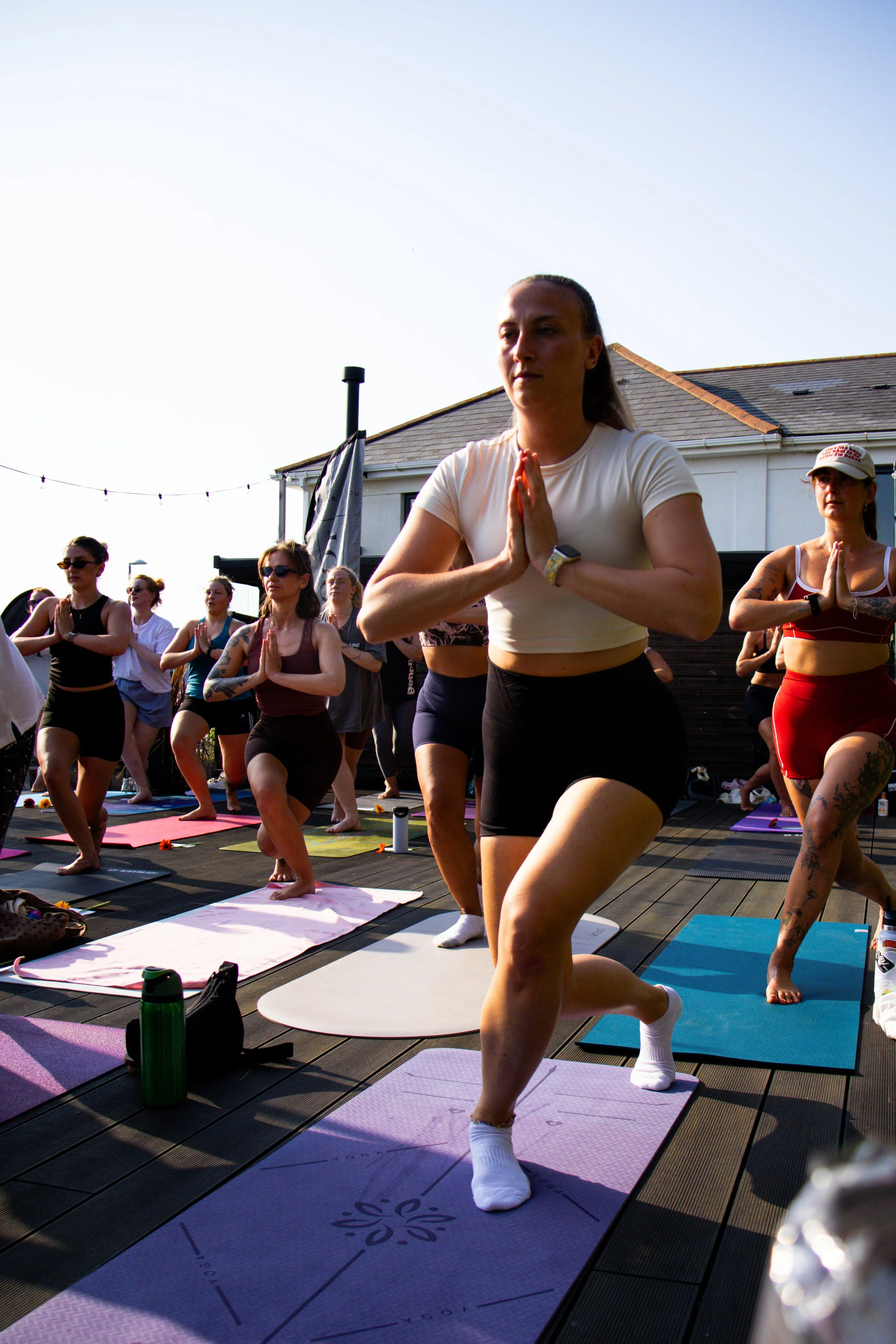 Group of women practicing pilates outdoors on a wooden deck, performing a kneeling prayer pose with hands in a namaste position, on yoga mats, under a clear sky.